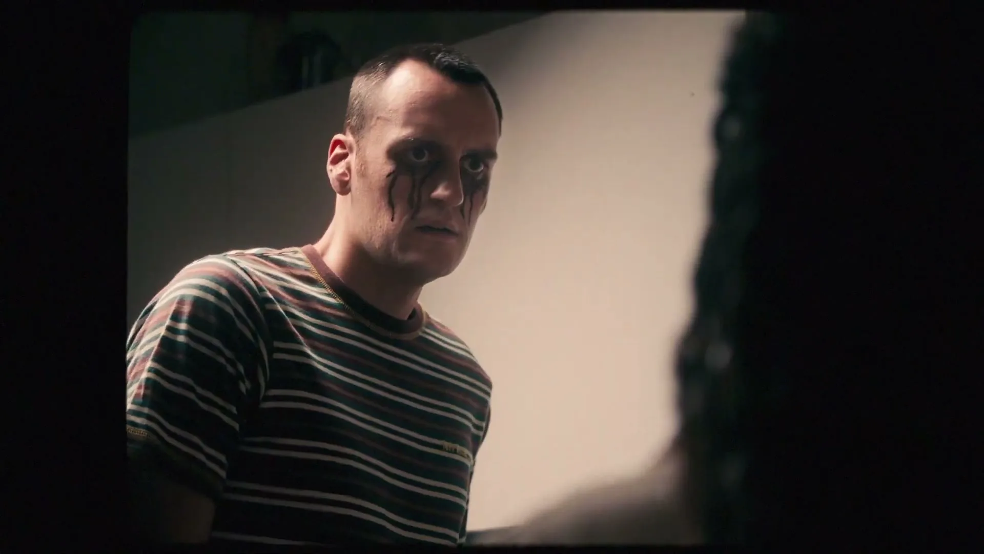 A man with black tears streaming down his face looks intensely at someone off-screen in a dimly lit room, appearing distressed.