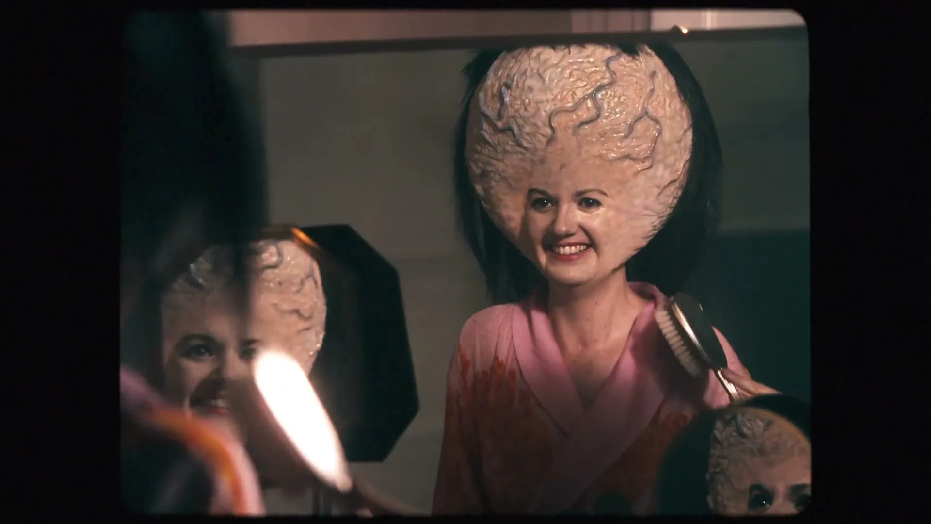 A woman with an enlarged, brain-like head, wearing a pink robe, brushes her hair in front of a mirror, laughing maniacally.