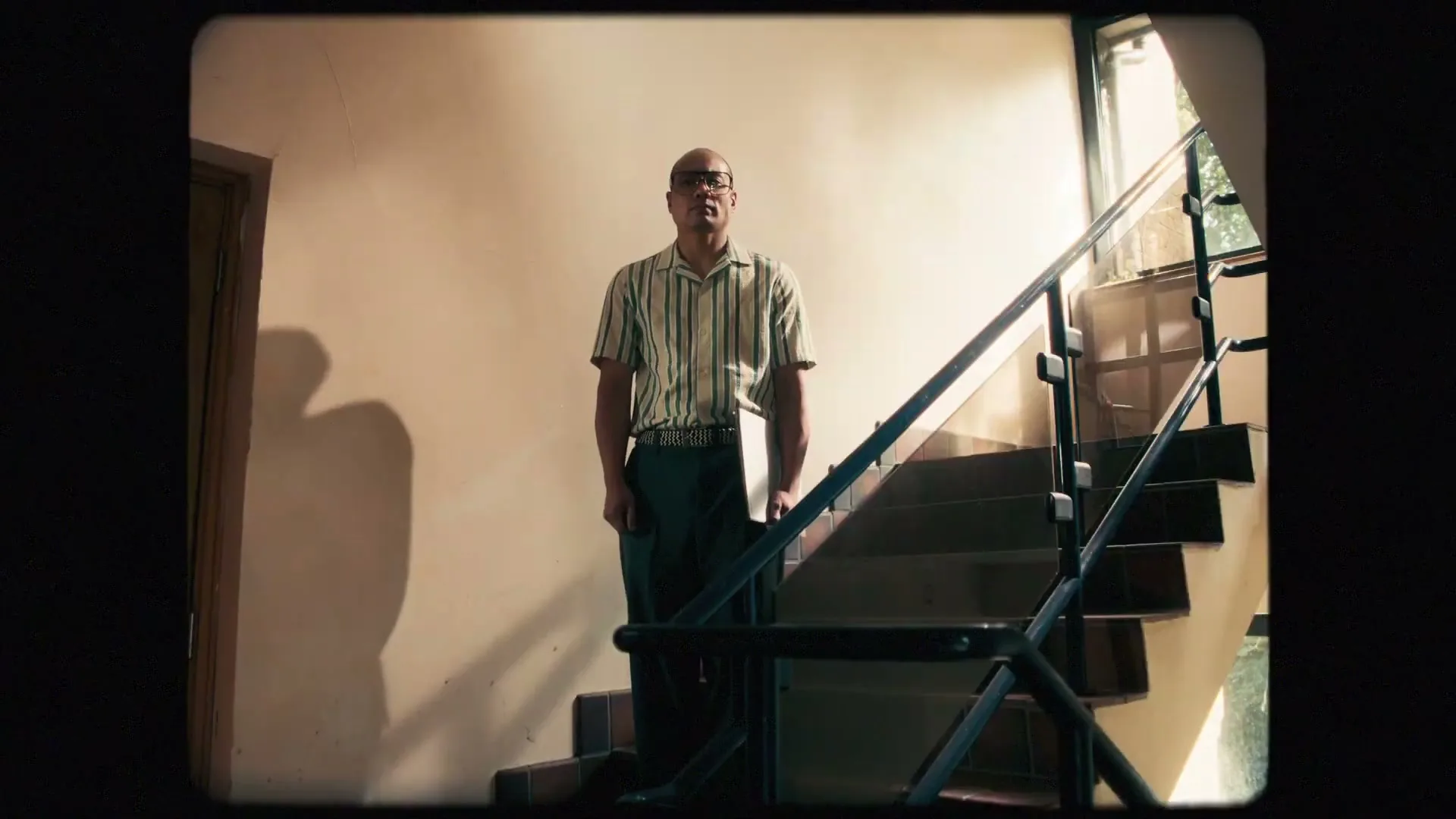 A man in his 30s, wearing a striped shirt and glasses, stands on a staircase as black ink splatters on the wall behind him.