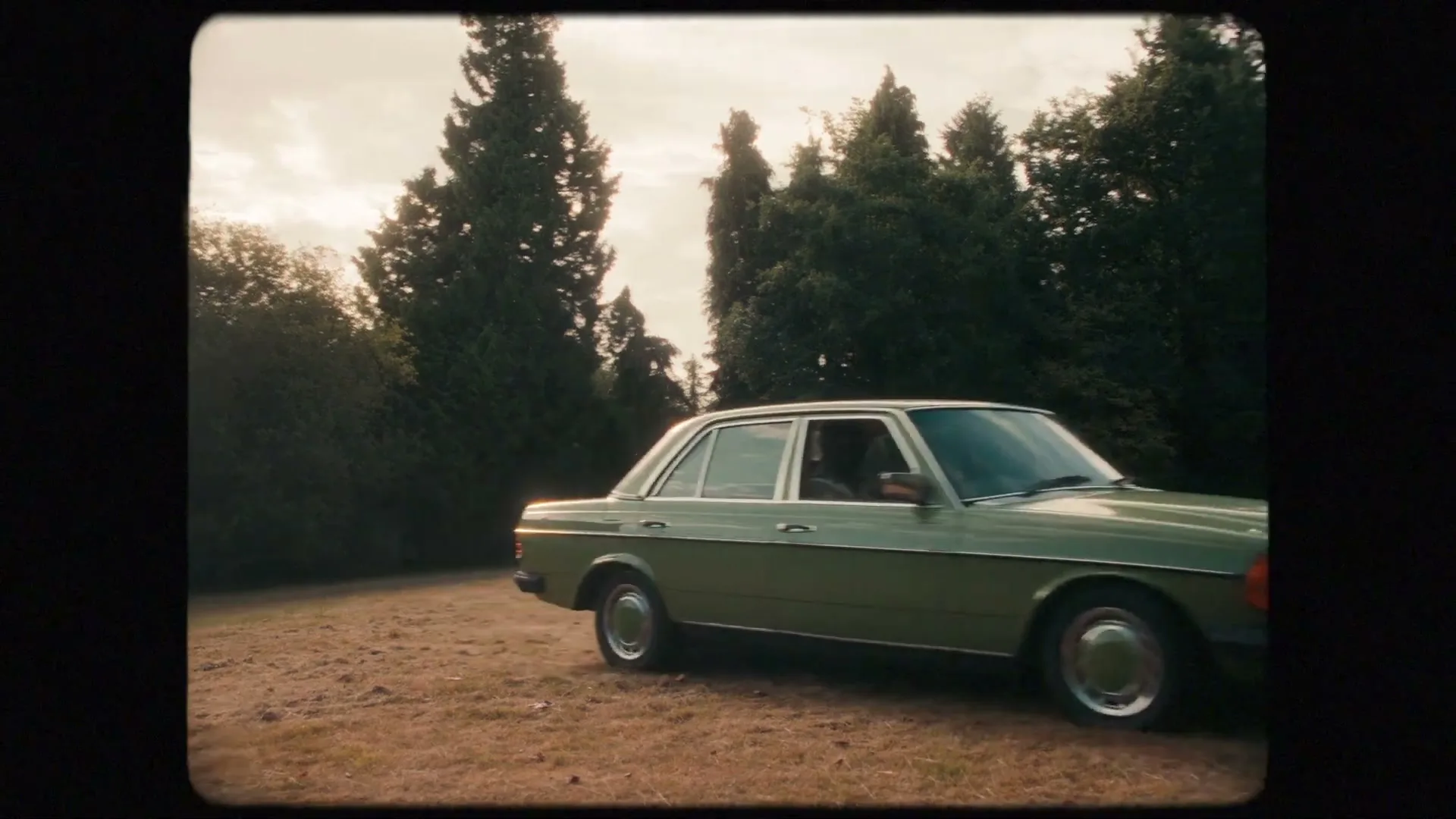 A vintage green car drives slowly across a dry, grassy field surrounded by tall trees under a cloudy sky.