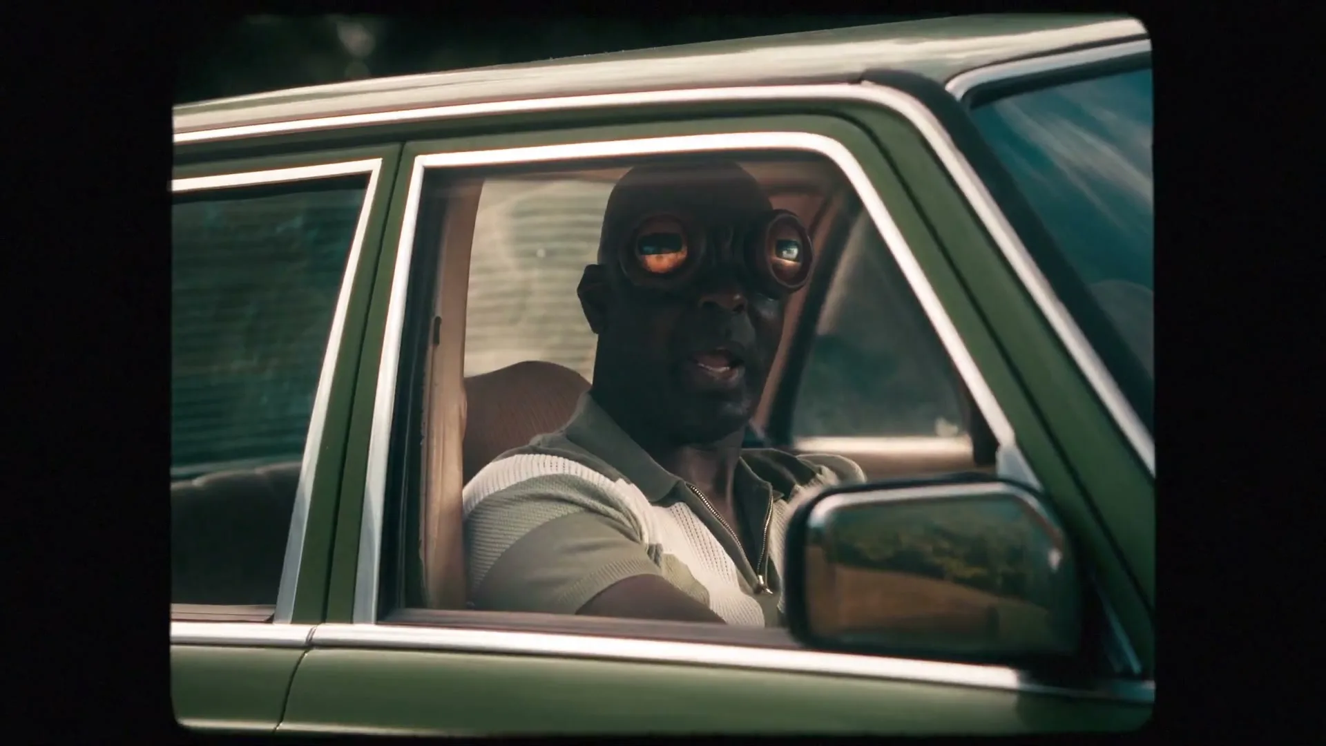 A man with large, round, reflective eyes sits in the driver's seat of a green car, looking forward with a distressed expression.