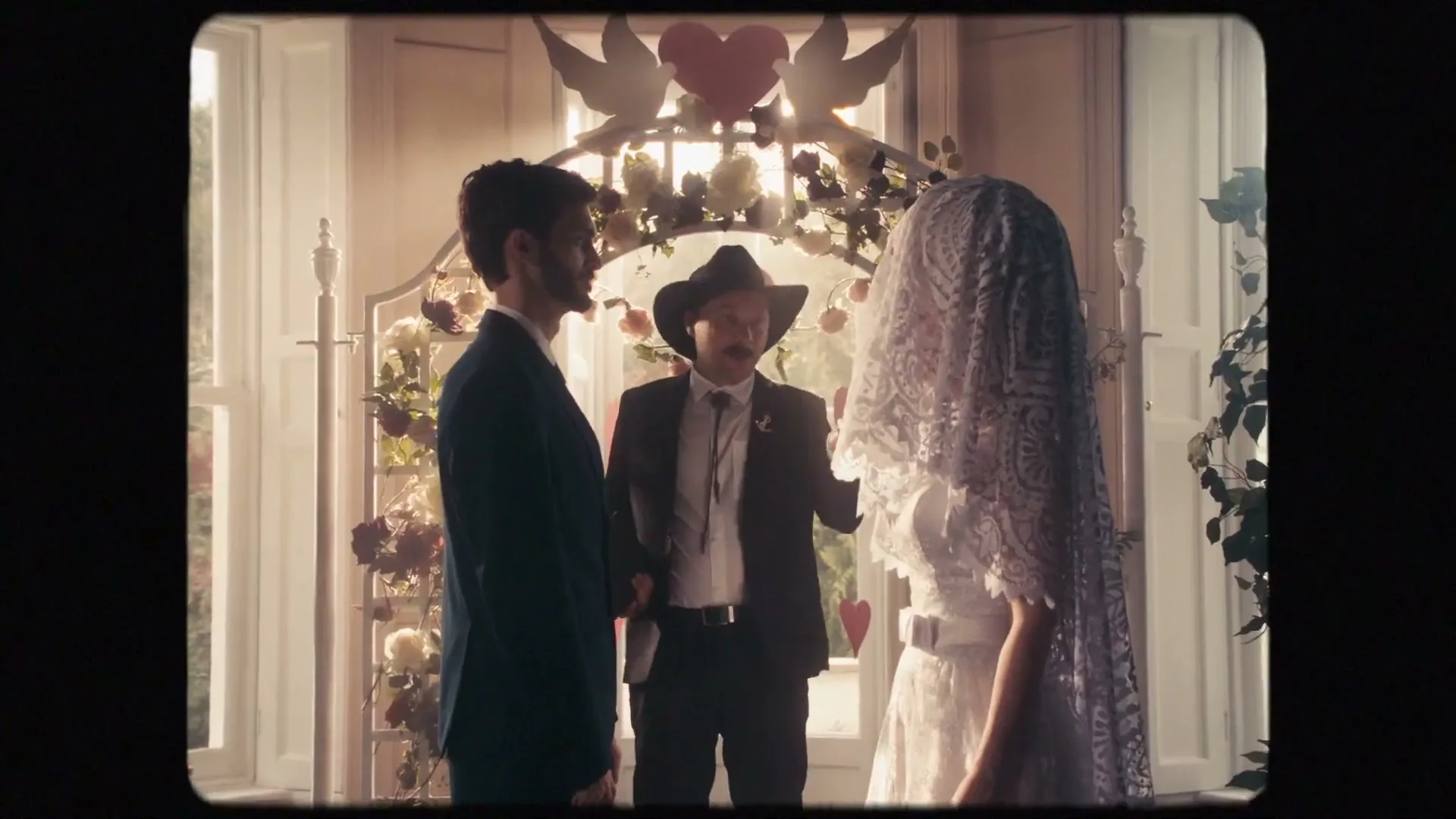 A man in a suit and a woman in a wedding dress stand before an officiant in a cowboy hat, under a floral archway in a bright room.