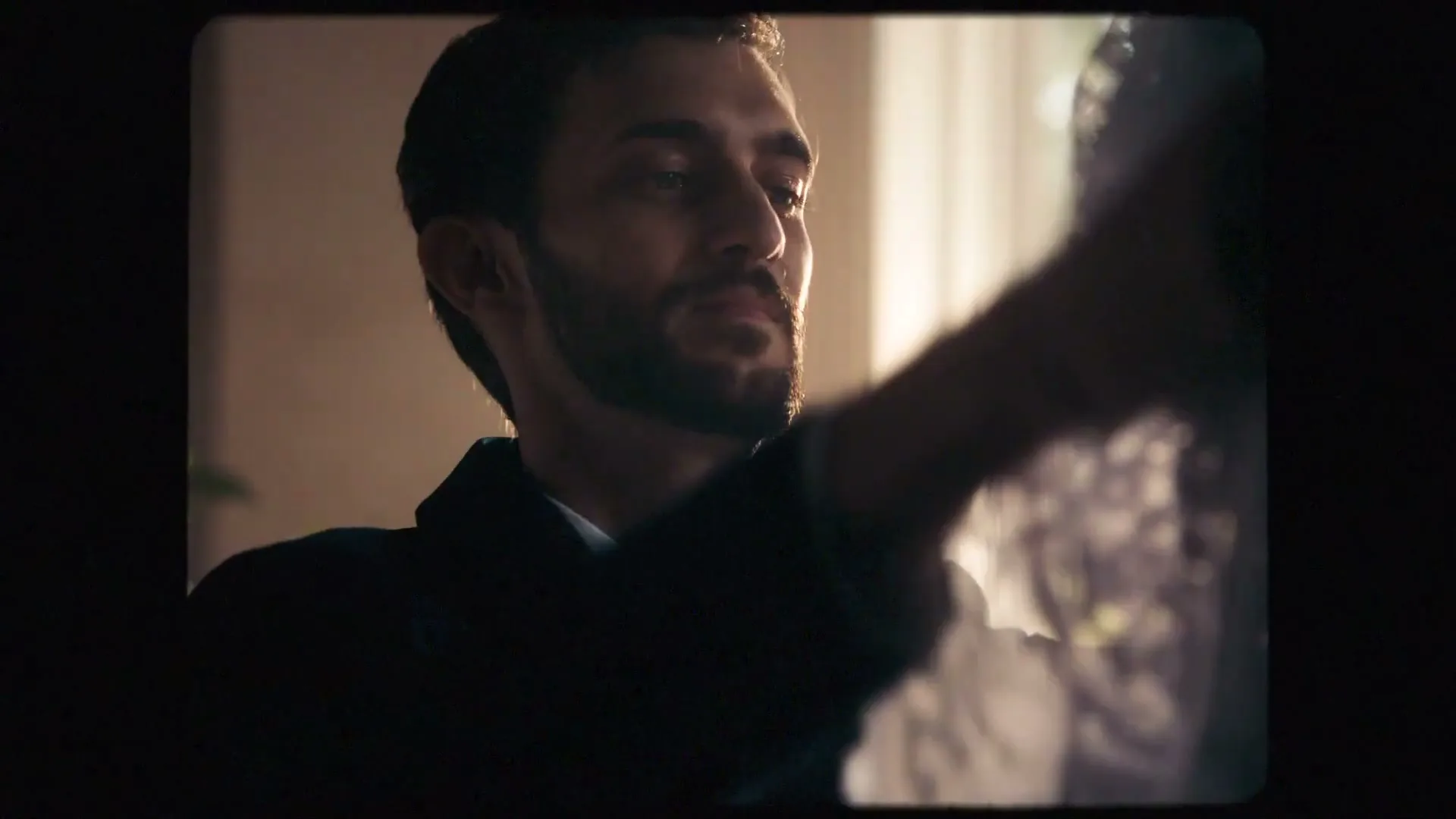 A man with a beard in a suit smiles at someone off-camera, with a window and curtains in the background.