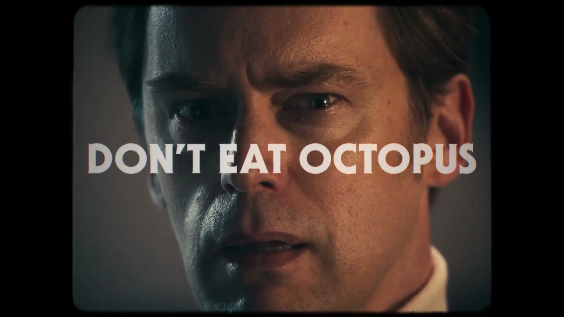 A man with a serious expression looks directly at the camera, with text overlaid on his face, in an interior setting.