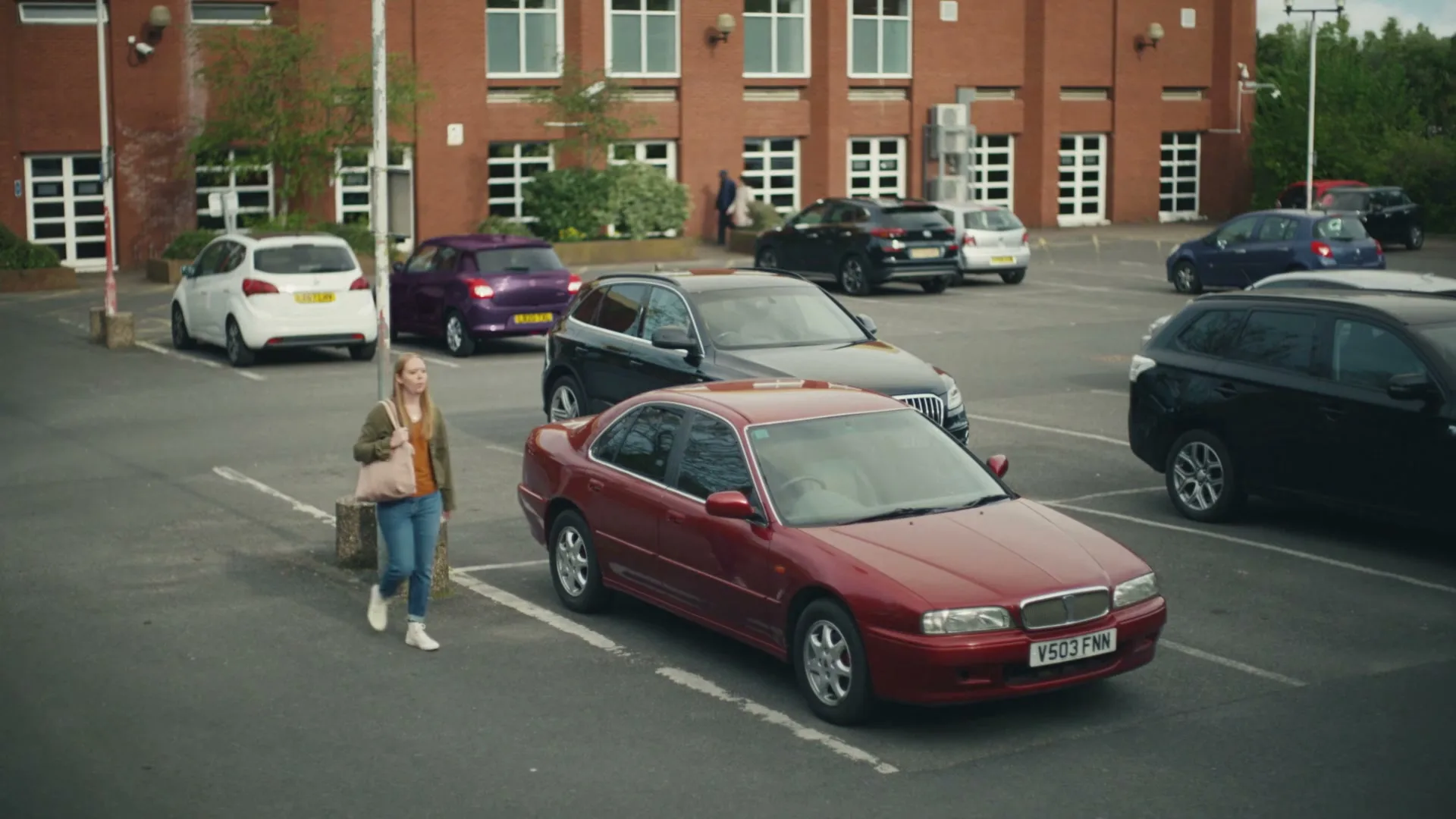 A young woman in a green jacket and jeans walks through a parking lot towards a red car, with a brick building in the background.
