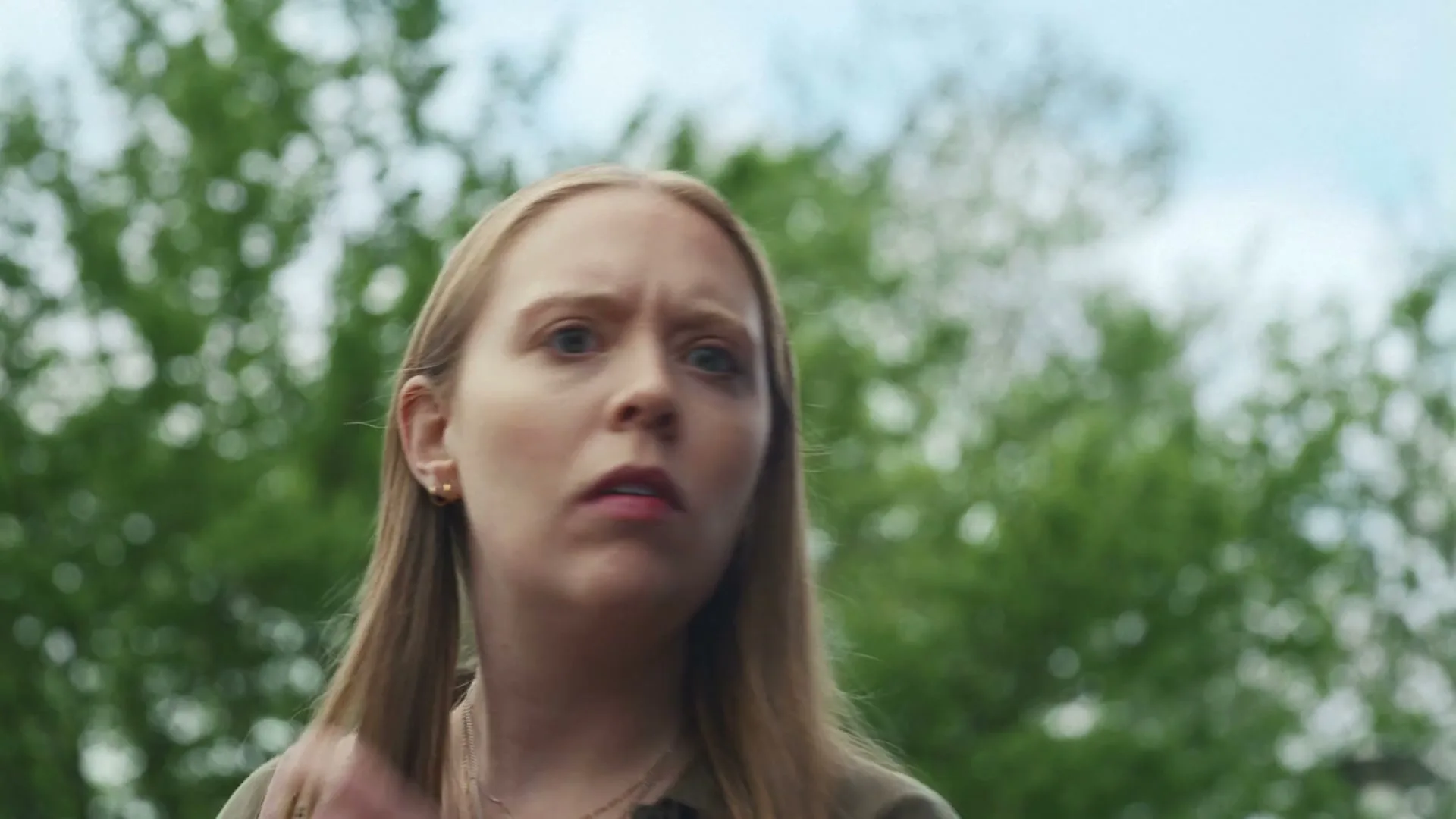 A young woman with long blonde hair looks up with a surprised expression, scratching her head, against a blurred background of green trees and blue sky.