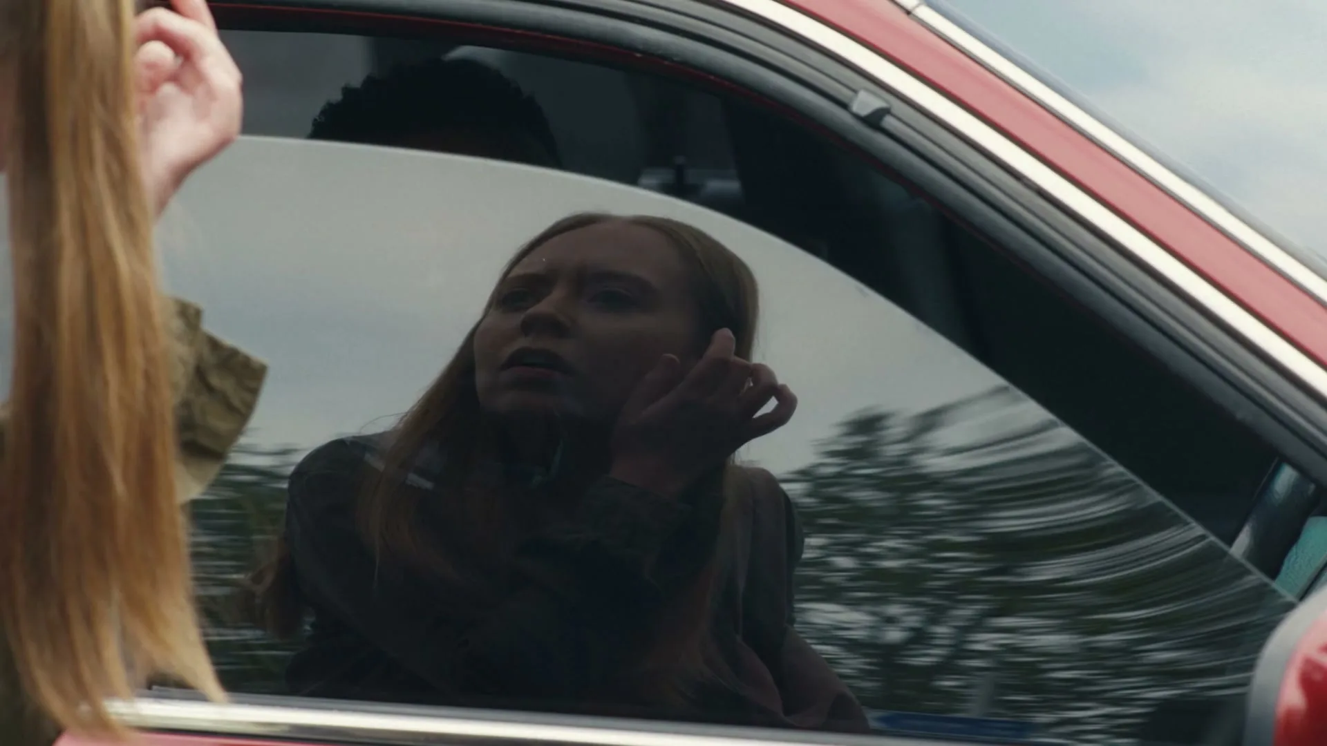A young boy with dark skin and a plaid shirt looks out of a car window, his reflection visible in the glass, as a woman with long blonde hair stands outside the car.