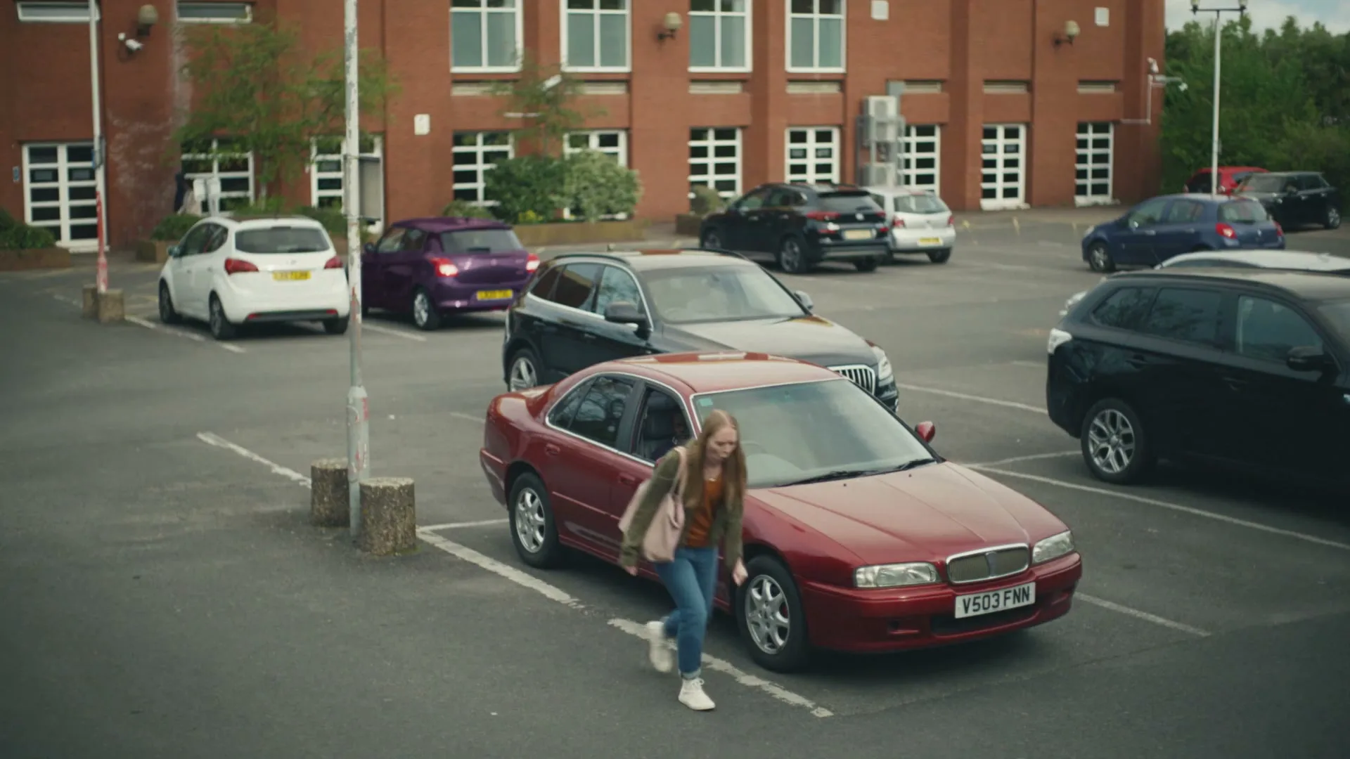 A young woman with long blonde hair and a backpack gets out of a red car in a parking lot in front of a brick building.