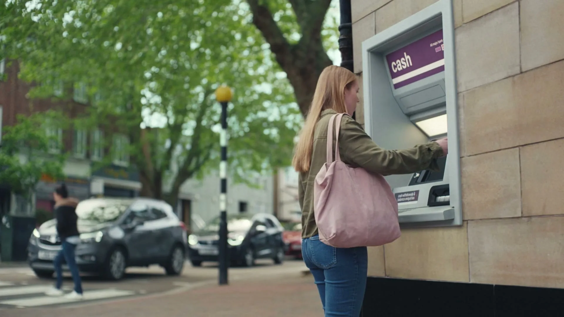 A young woman with long blonde hair and a pink tote bag uses an ATM on a city street during the day.