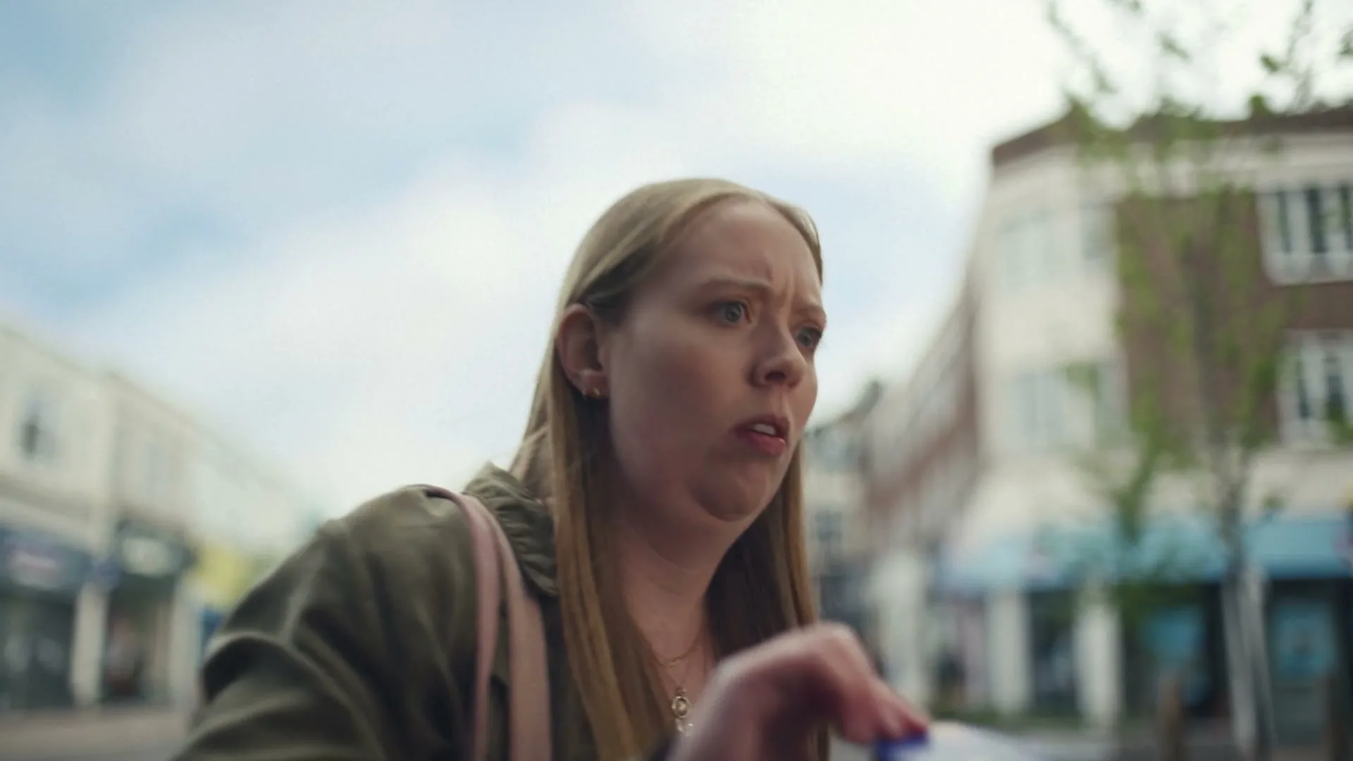 A young woman with long blonde hair, wearing a green jacket, looks confused while holding a small blue object on a street with shops in the background.