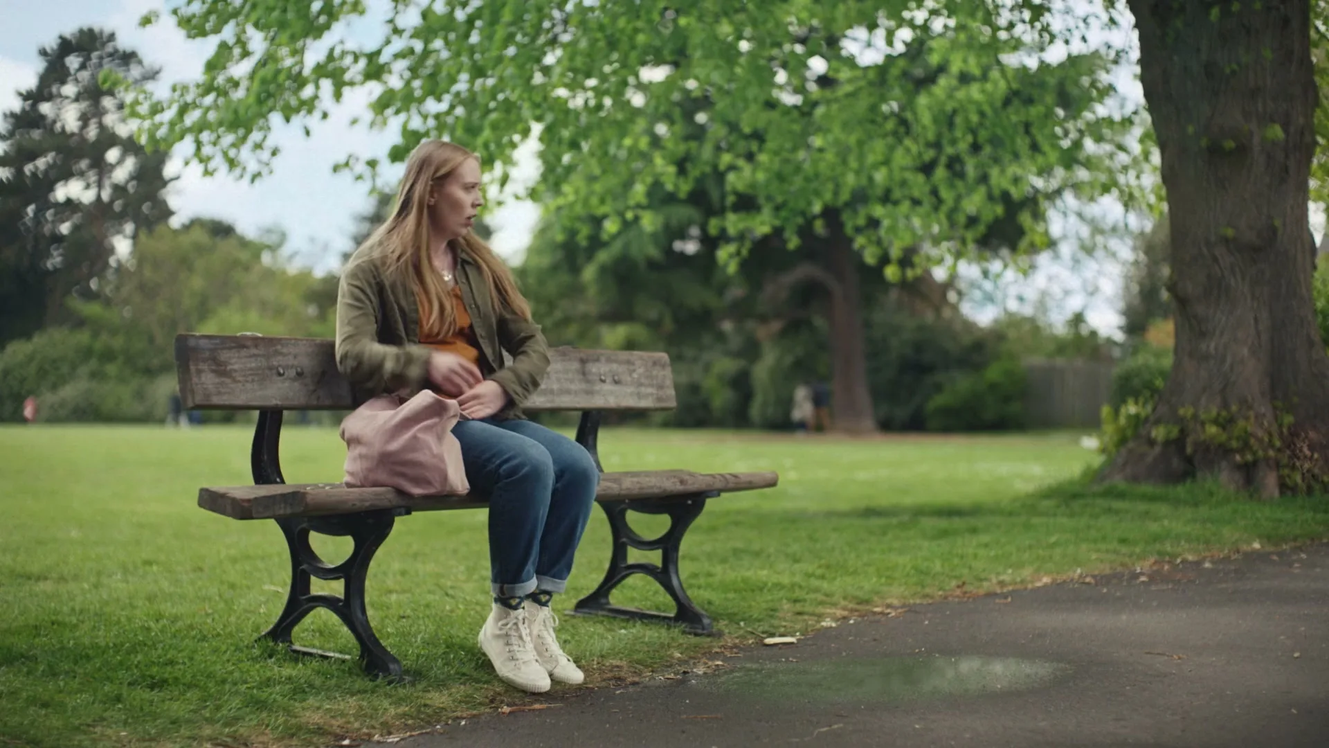 A young woman with long blonde hair, wearing a green jacket and jeans, sits down on a park bench, looking tired.