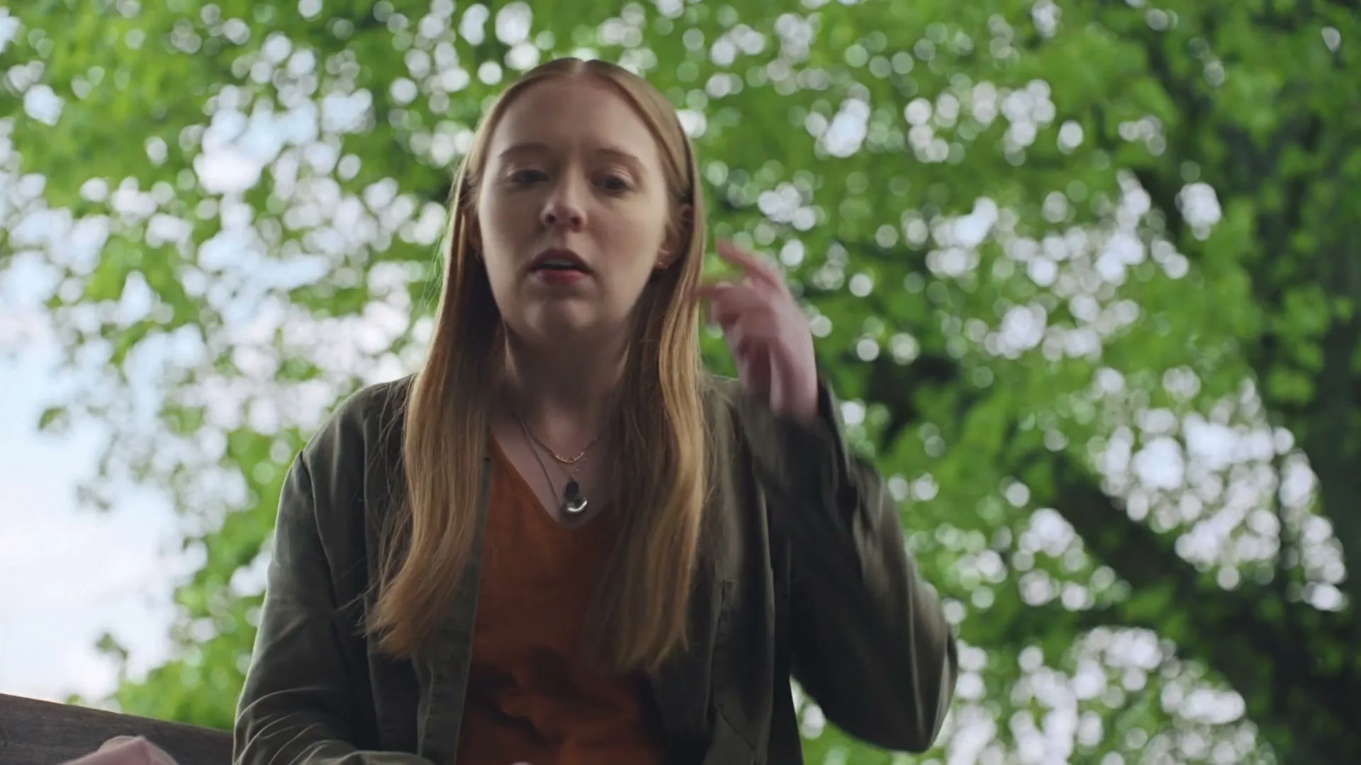 A young woman with long red hair, wearing a green jacket and orange top, sits outdoors and touches her hair, with green leaves in the background.