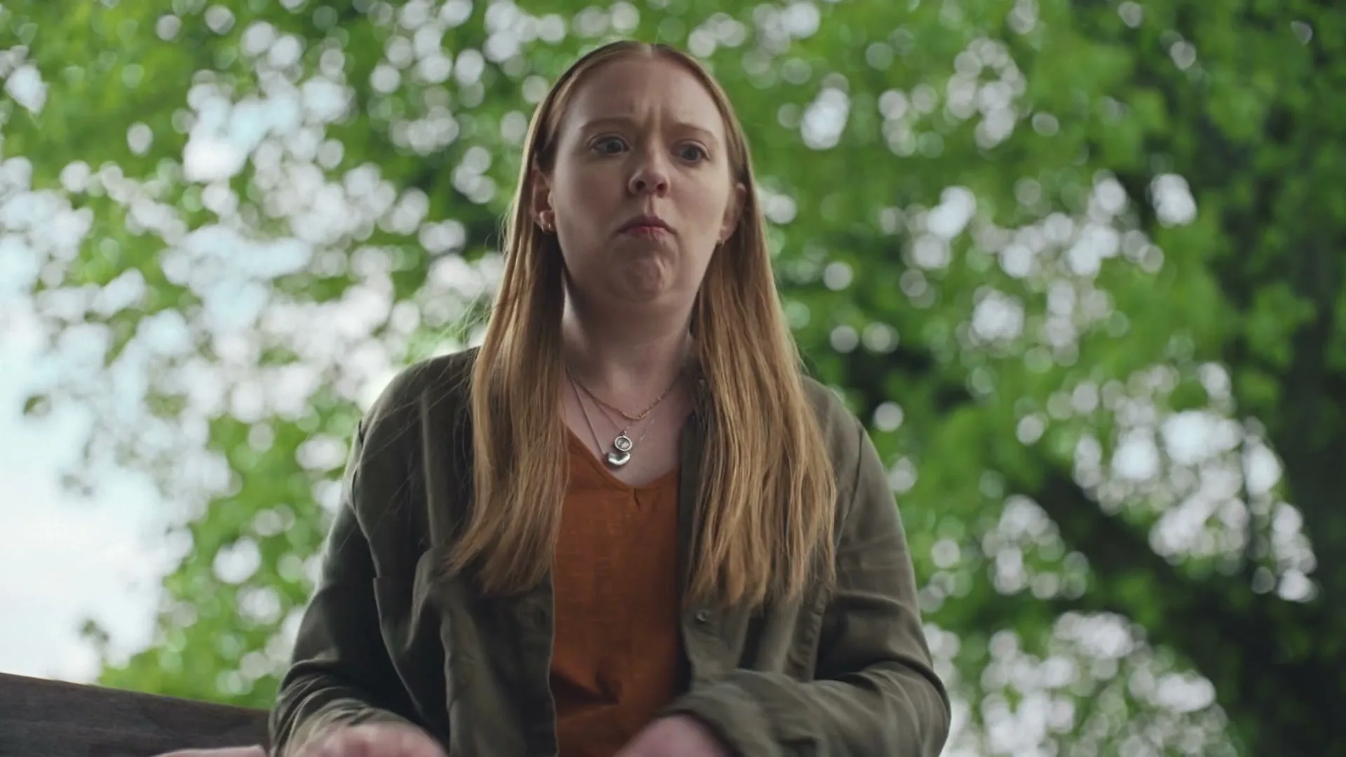 A young woman with long red hair, wearing a green jacket and orange top, looks down with a concerned expression in an outdoor setting with green trees in the background.