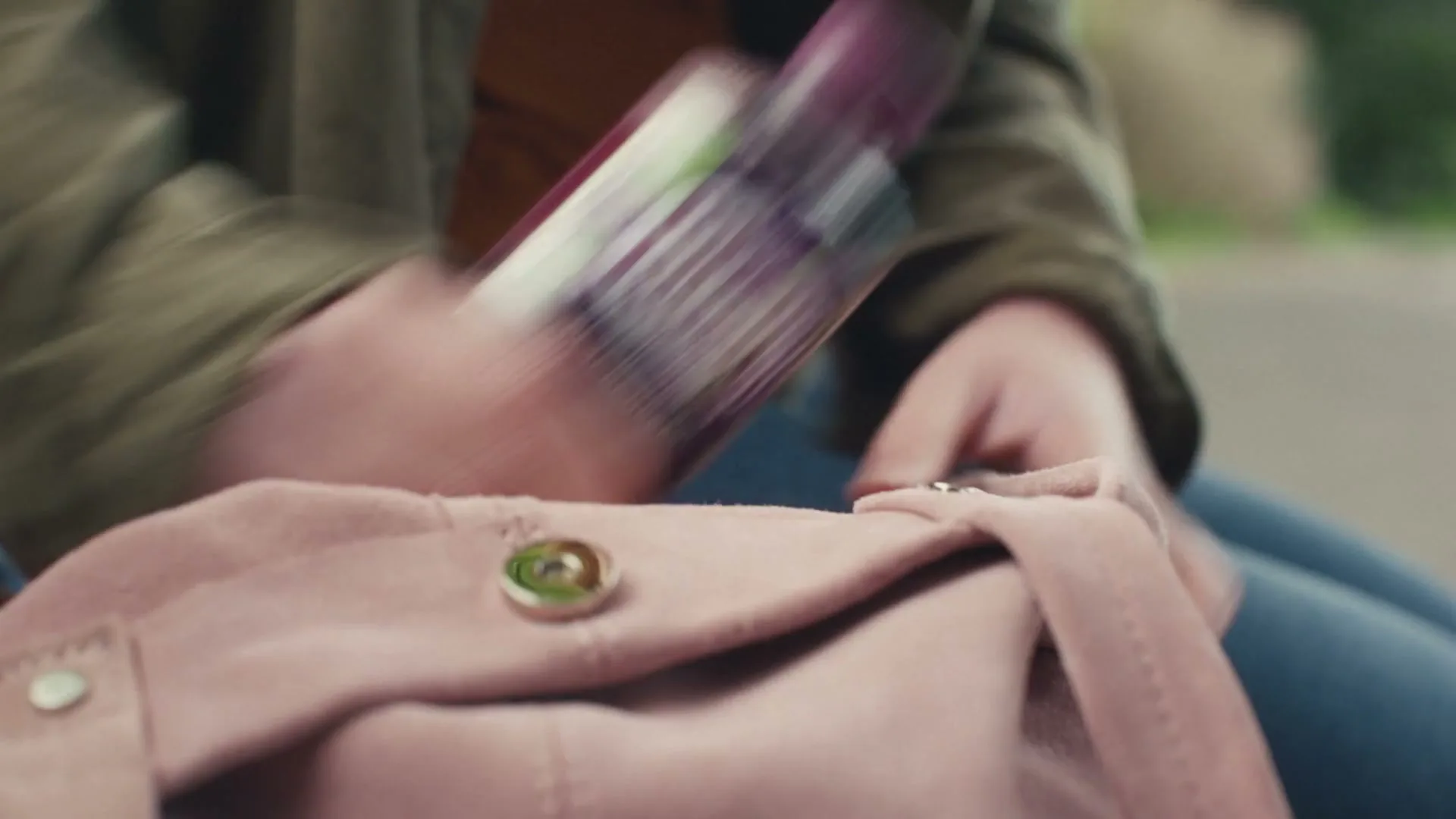 A person in a green jacket and blue jeans reaches into a pink suede bag to pull out a Ribena bottle, outdoors.