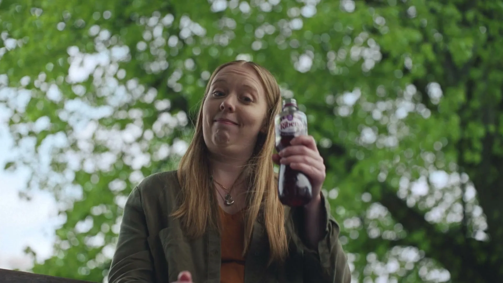 A young woman with long red hair, wearing a green jacket and orange top, holds up a bottle of Ribena and smiles at the camera, outdoors under green trees.