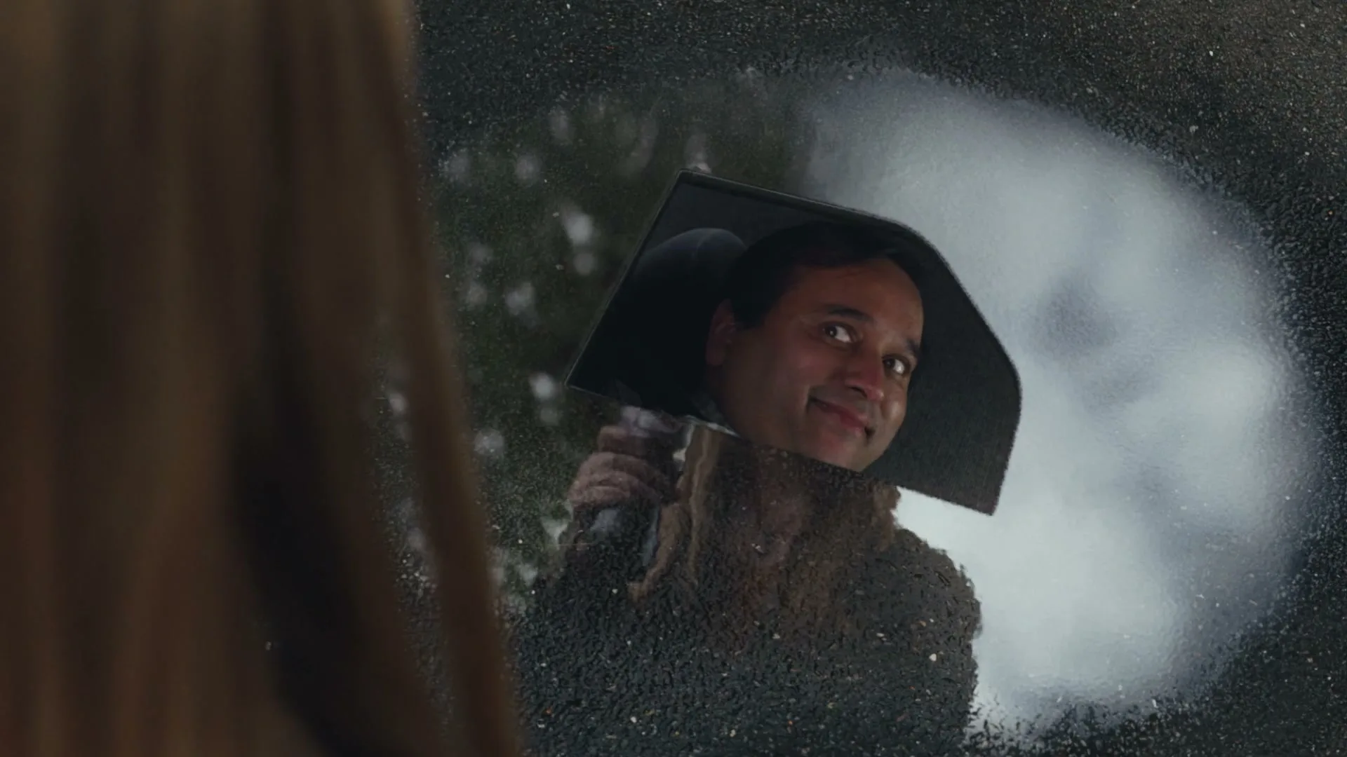 A man's face is reflected in a puddle on the ground, smiling, as a woman's blurry shoulder is in the foreground.