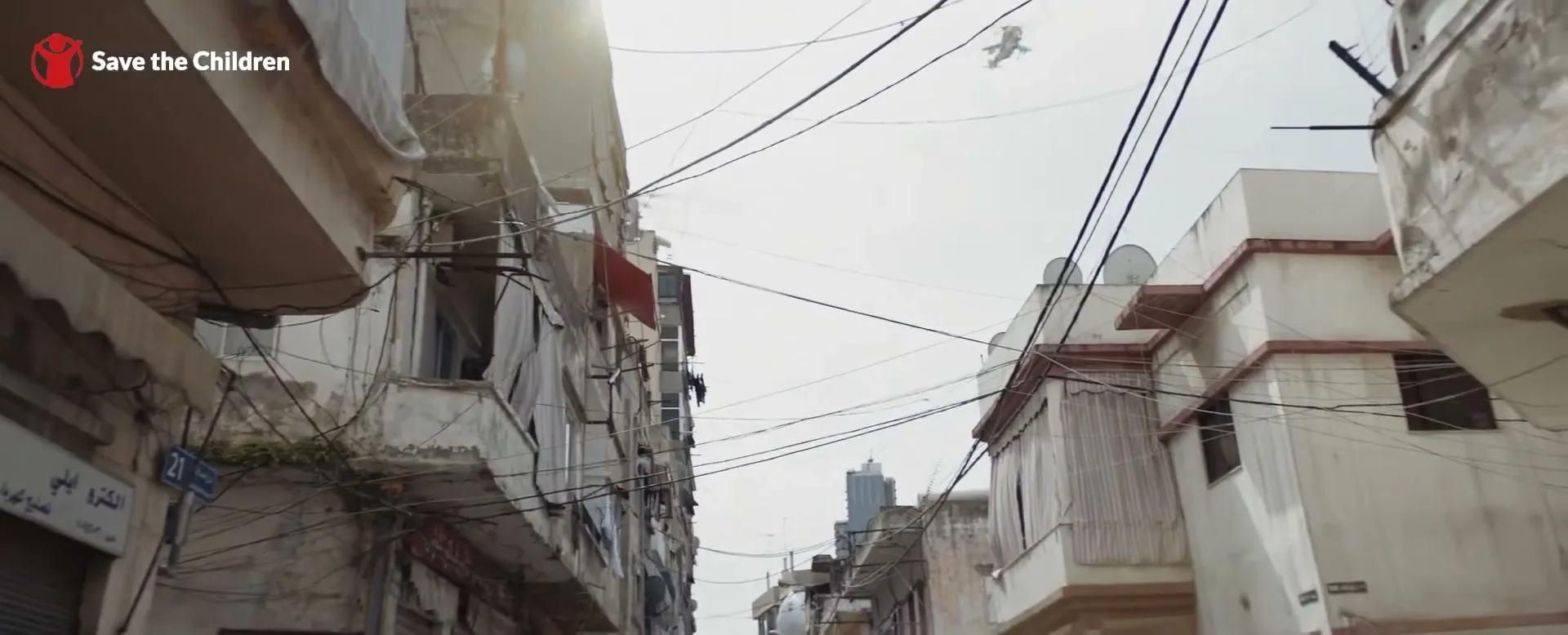 A low-angle shot captures a military helicopter flying over a narrow street lined with old, worn buildings and tangled power lines under a bright sky.