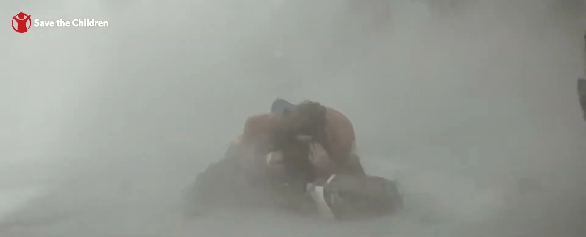 A person in a red jacket shields a child on the ground amidst a cloud of dust and debris, likely after an explosion or collapse.