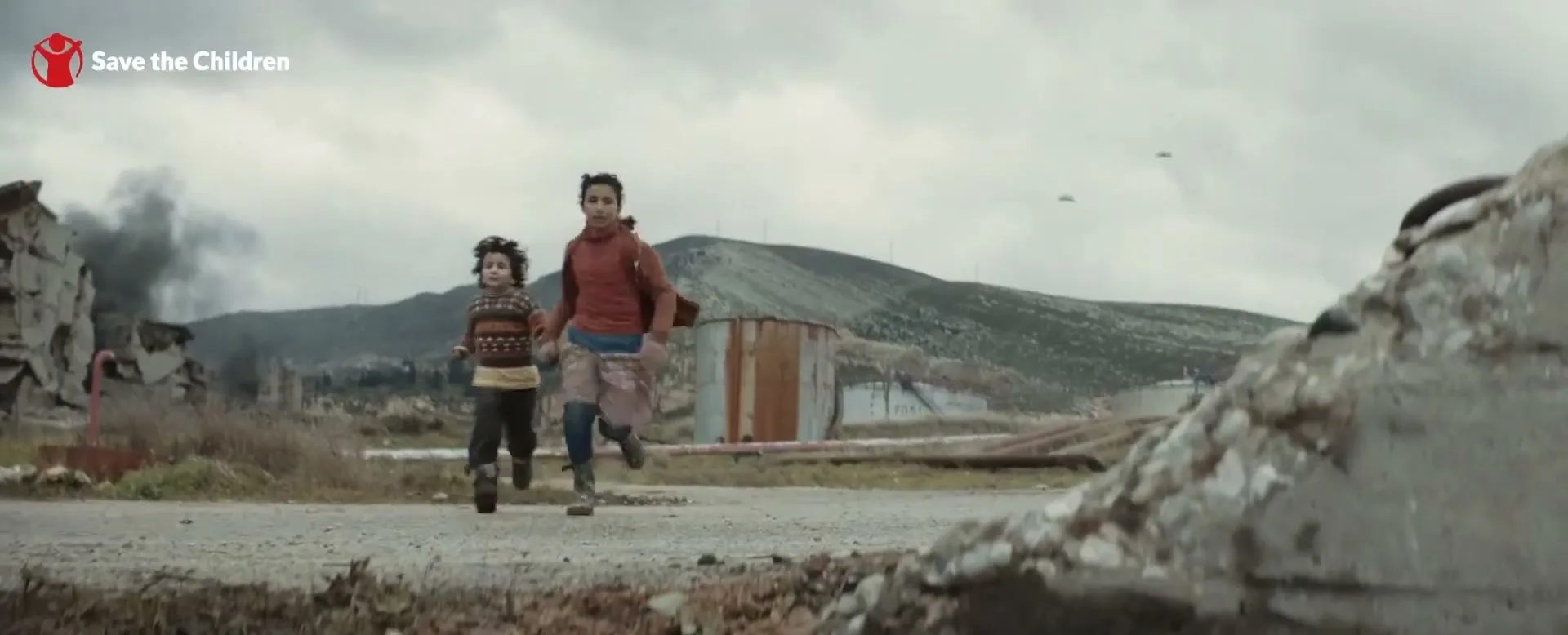 Two children, a young girl and a boy, run hand-in-hand down a dirt road in a war-torn landscape with smoke rising in the background.