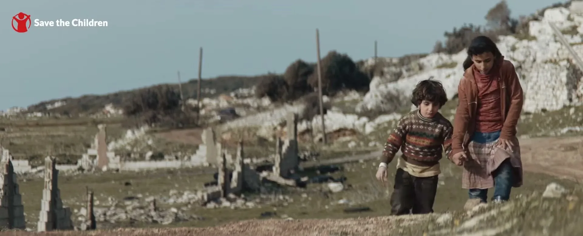 A young girl and a boy walk hand-in-hand through a desolate, war-torn landscape under a clear blue sky, with ruined buildings in the background.