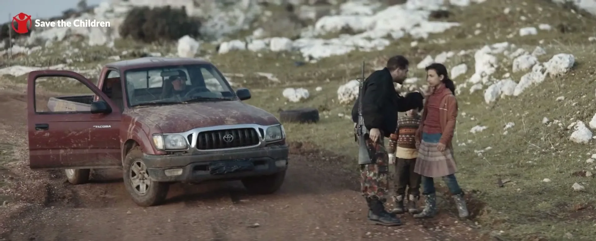 A man in military fatigues talks to a young girl and boy next to a red pickup truck on a dirt road in a rocky, grassy landscape.