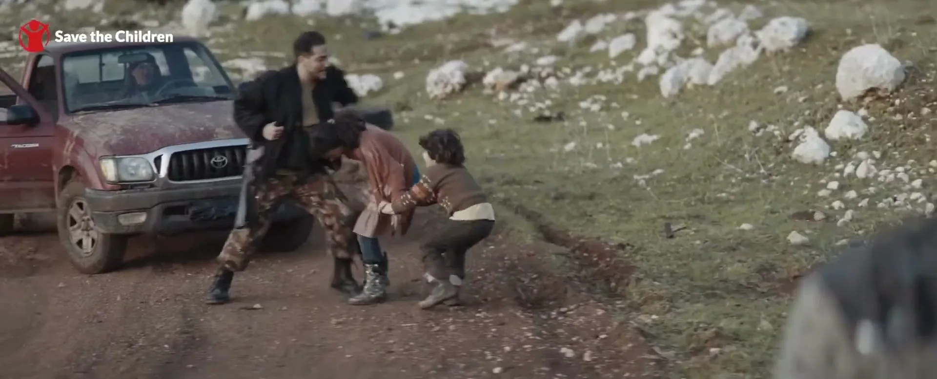 A man in military fatigues struggles with two children on a dirt road next to a red pickup truck, with a blurred person in the foreground.