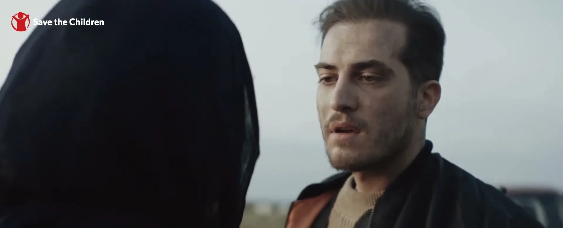A man with a beard and dirty face looks at someone off-screen, wearing a black jacket and a beige sweater, outdoors.