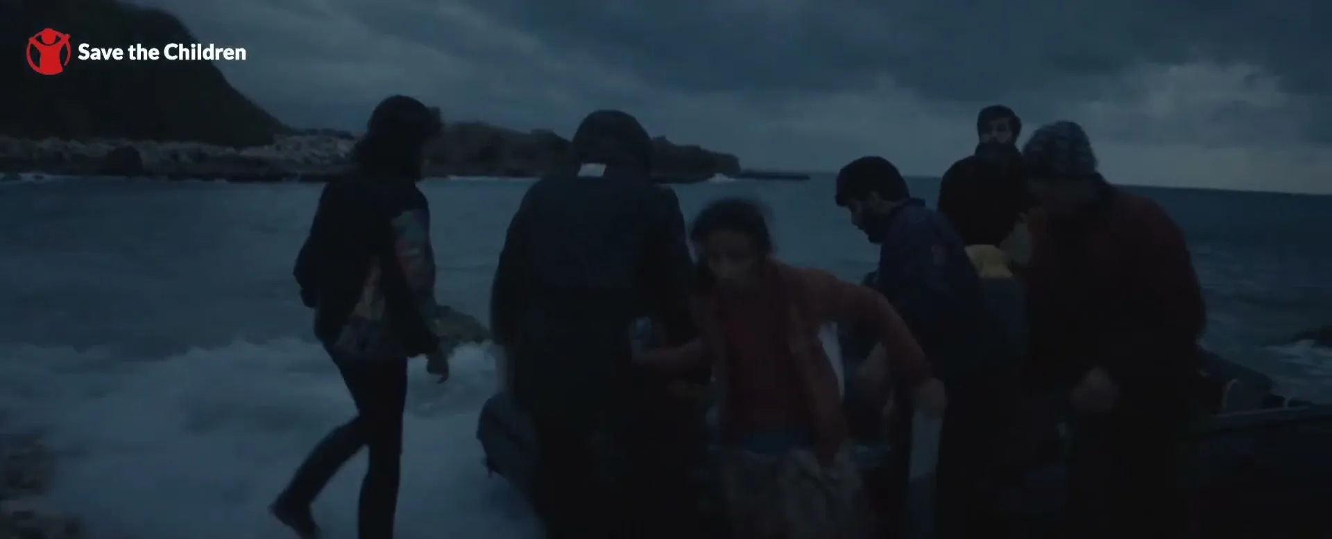 A group of refugees, including a child, disembark from a small boat onto a rocky shore under a dark, cloudy sky at dusk.