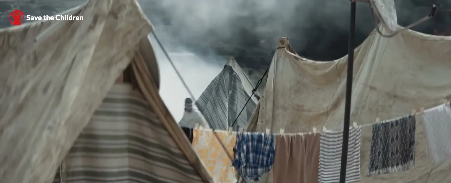 A wide shot shows a refugee camp with clothes hanging on a line in the foreground, and people moving between tents in the background under a smoky sky.