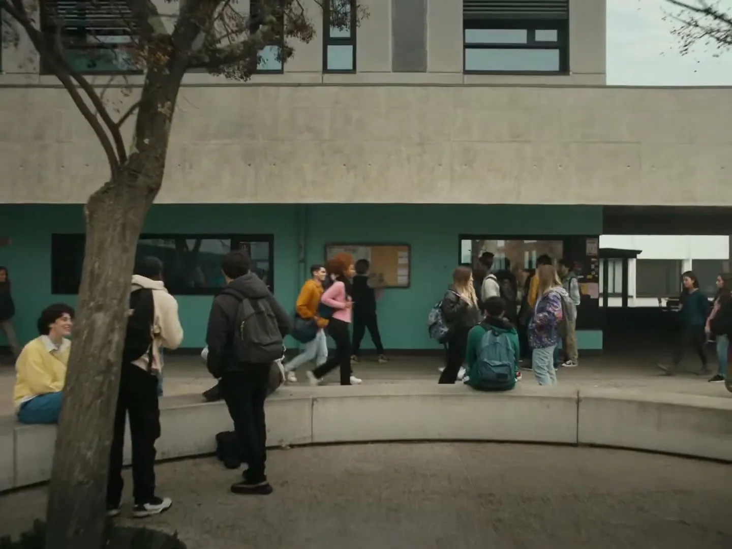 A diverse group of high school students walk and gather in a concrete courtyard outside a modern school building during the day.