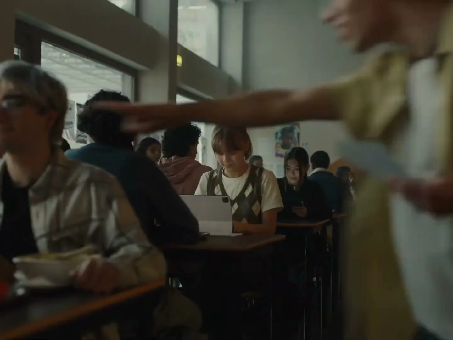 A male student in a yellow shirt walks past tables in a busy school cafeteria, pointing towards a female student using a tablet.