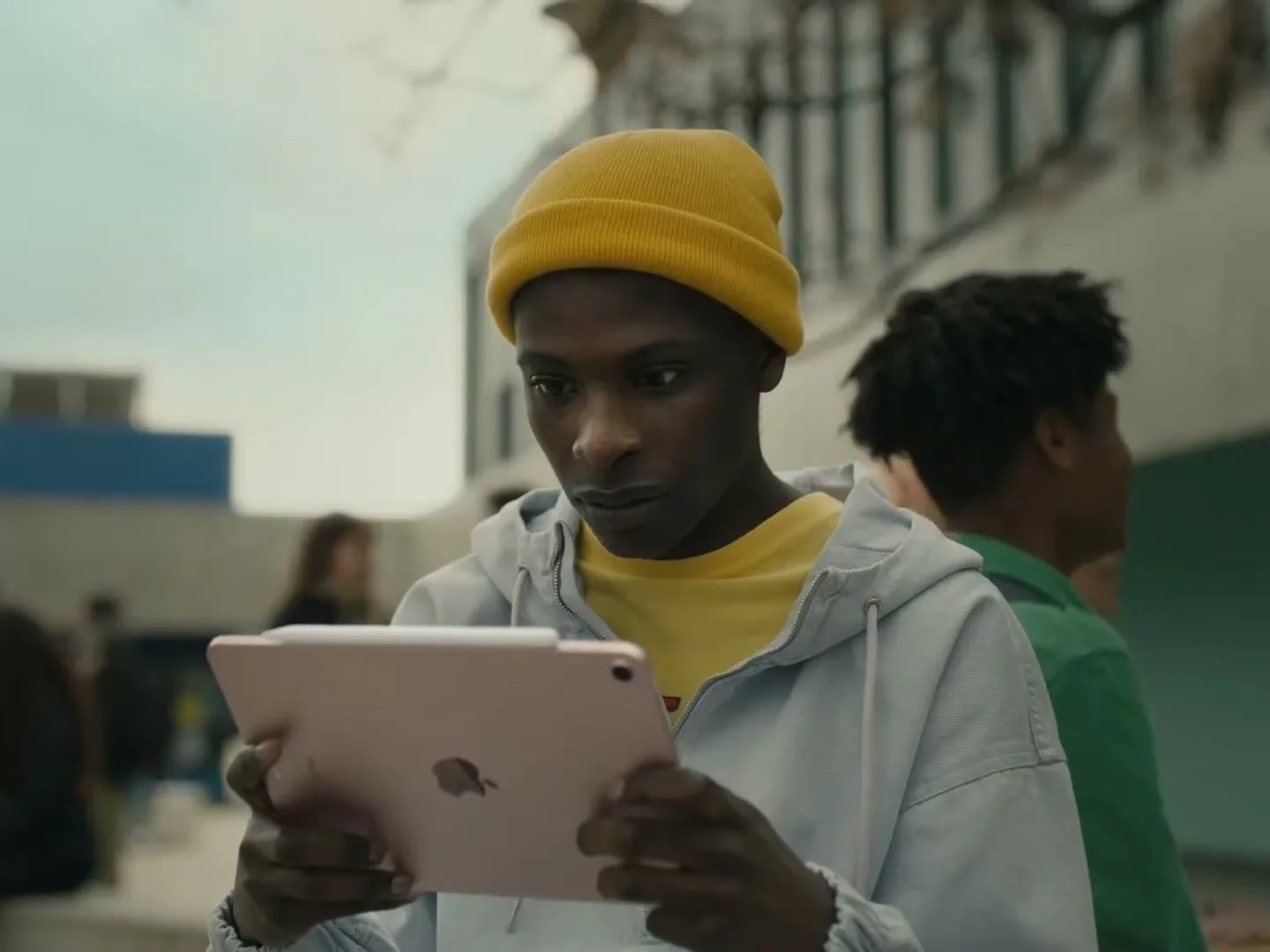 A young Black man in a yellow beanie and light blue hoodie looks intently at a pink iPad he holds in his hands, outdoors during the day.