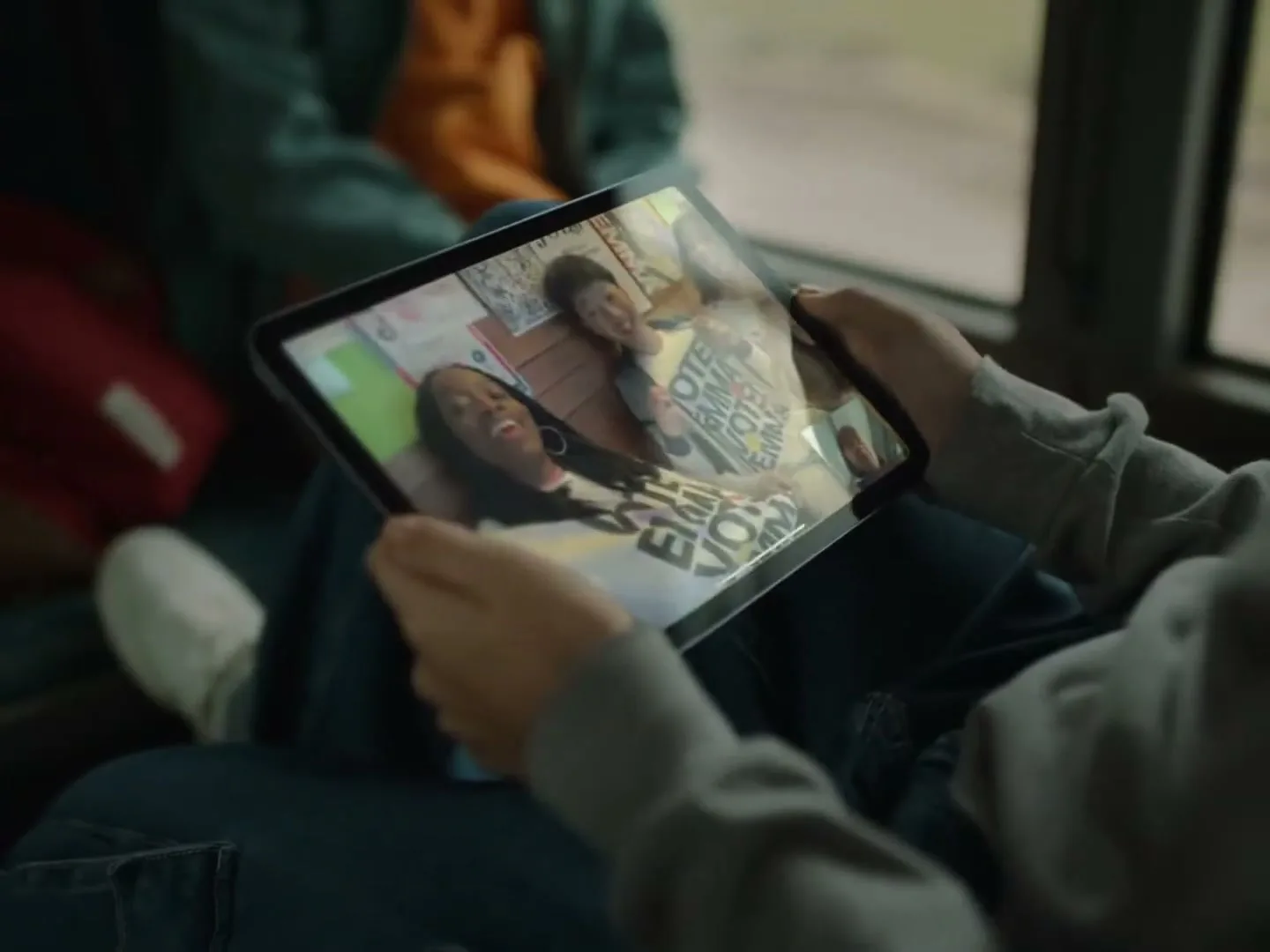 A person holds an iPad, displaying a video call with two young women laughing, while sitting on a bus.