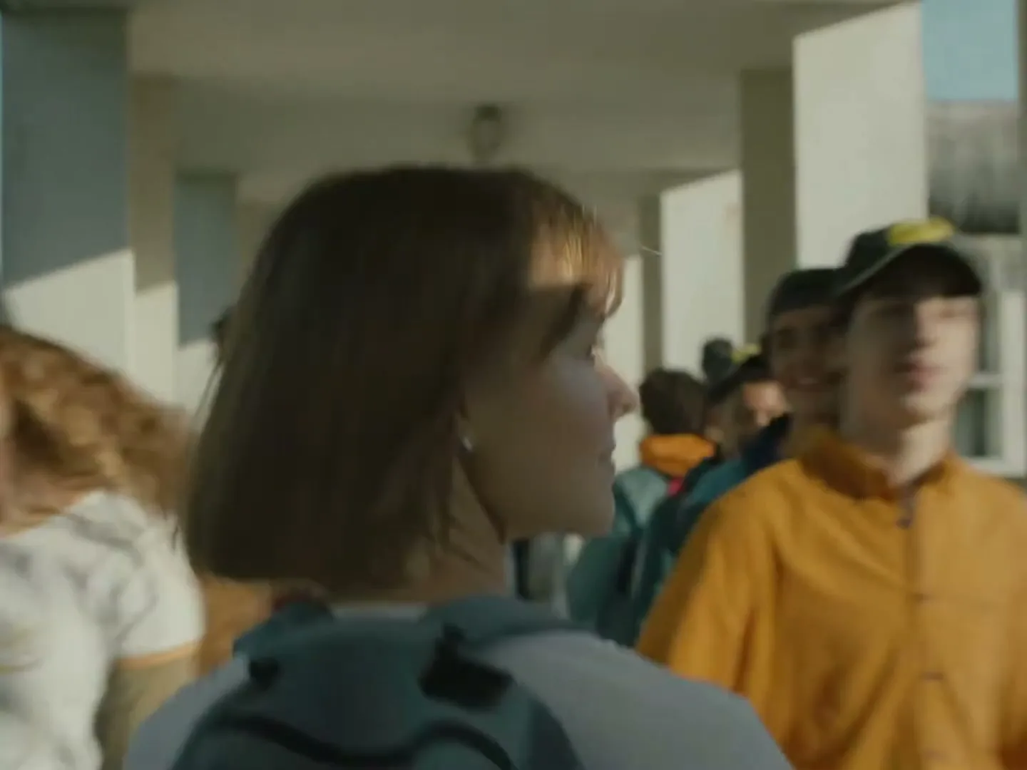 A young woman with short brown hair and a backpack walks through a crowded school hallway, turning her head to the side.