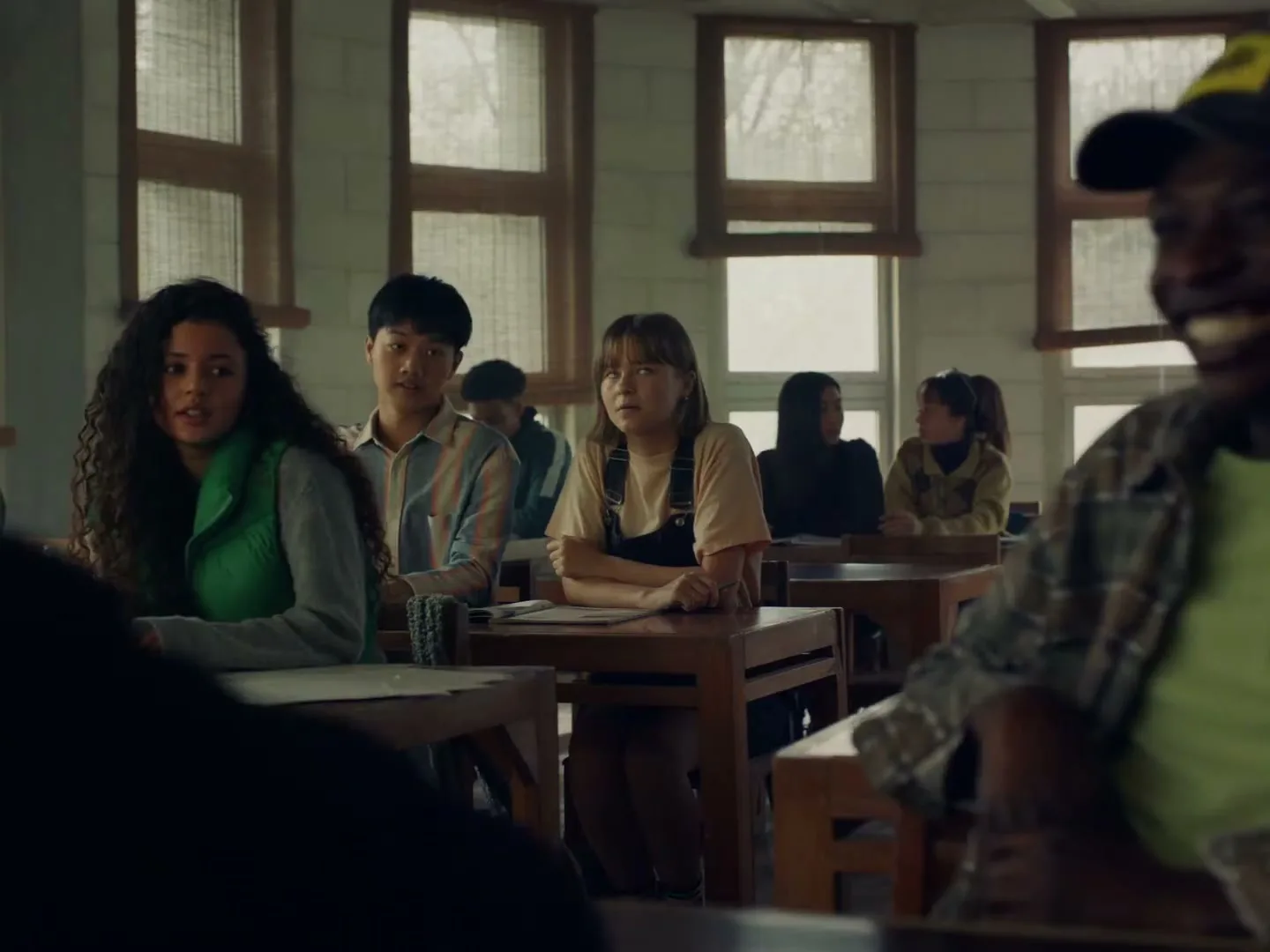 A diverse group of high school students sits at wooden desks in a classroom, looking towards the front with varied expressions.