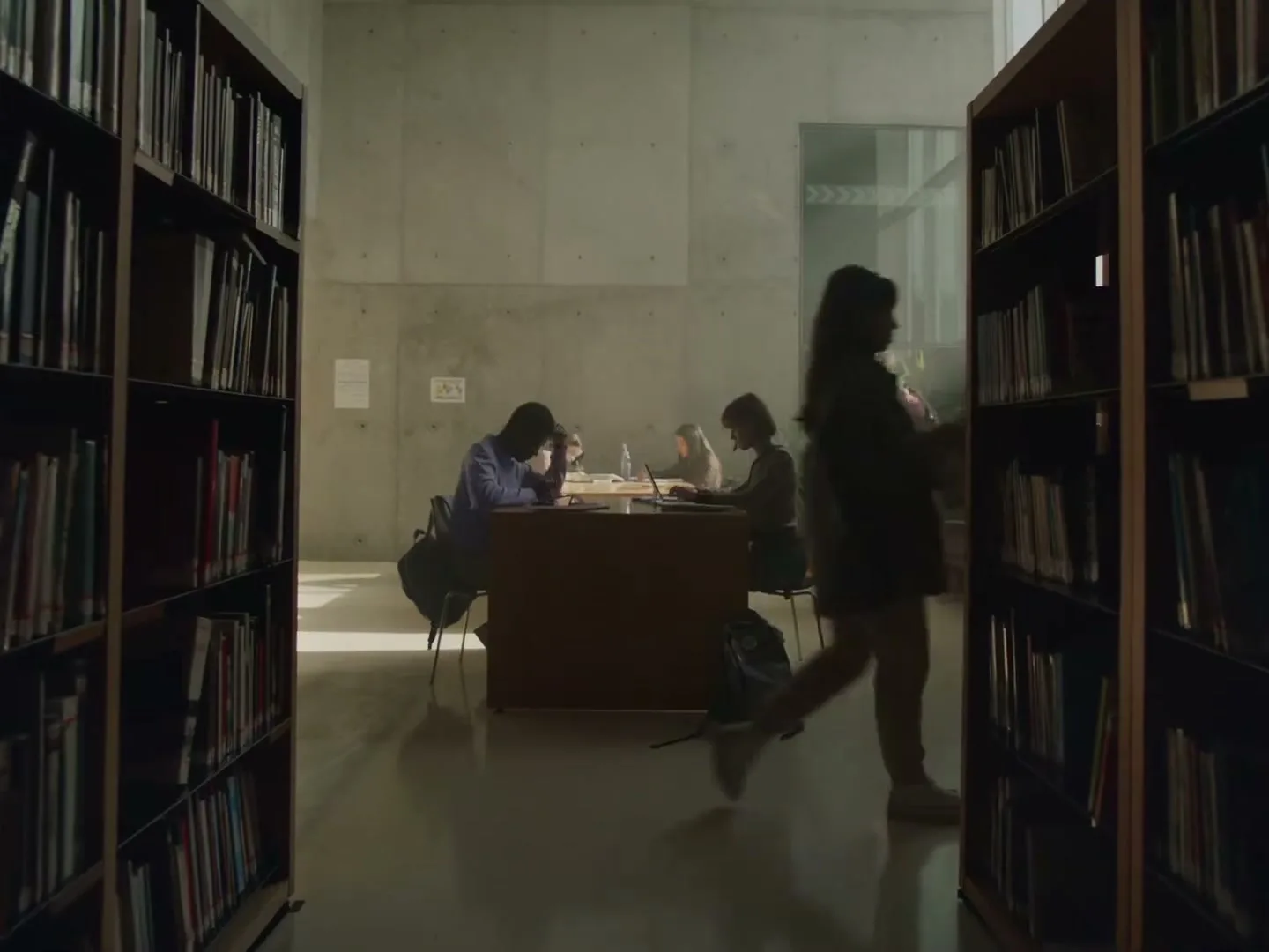 A young woman walks through a library aisle, passing students studying at a table, with sunlight streaming in from a window.