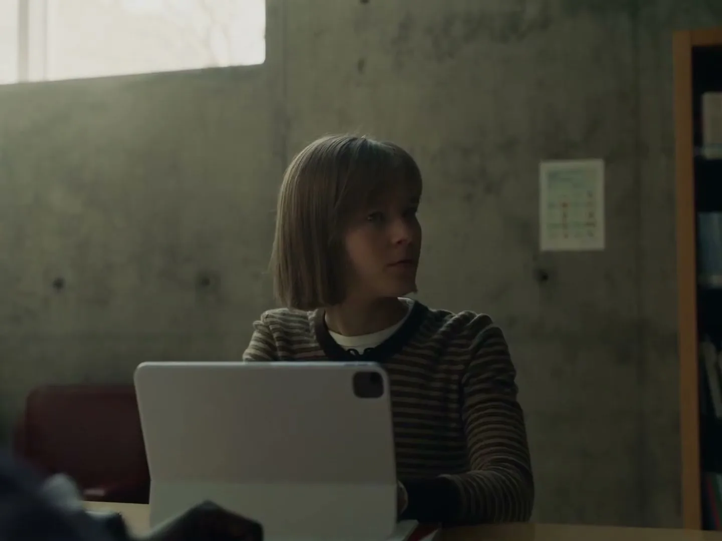 A young female student with a bob haircut sits at a desk with an iPad, looking to her right in a library.