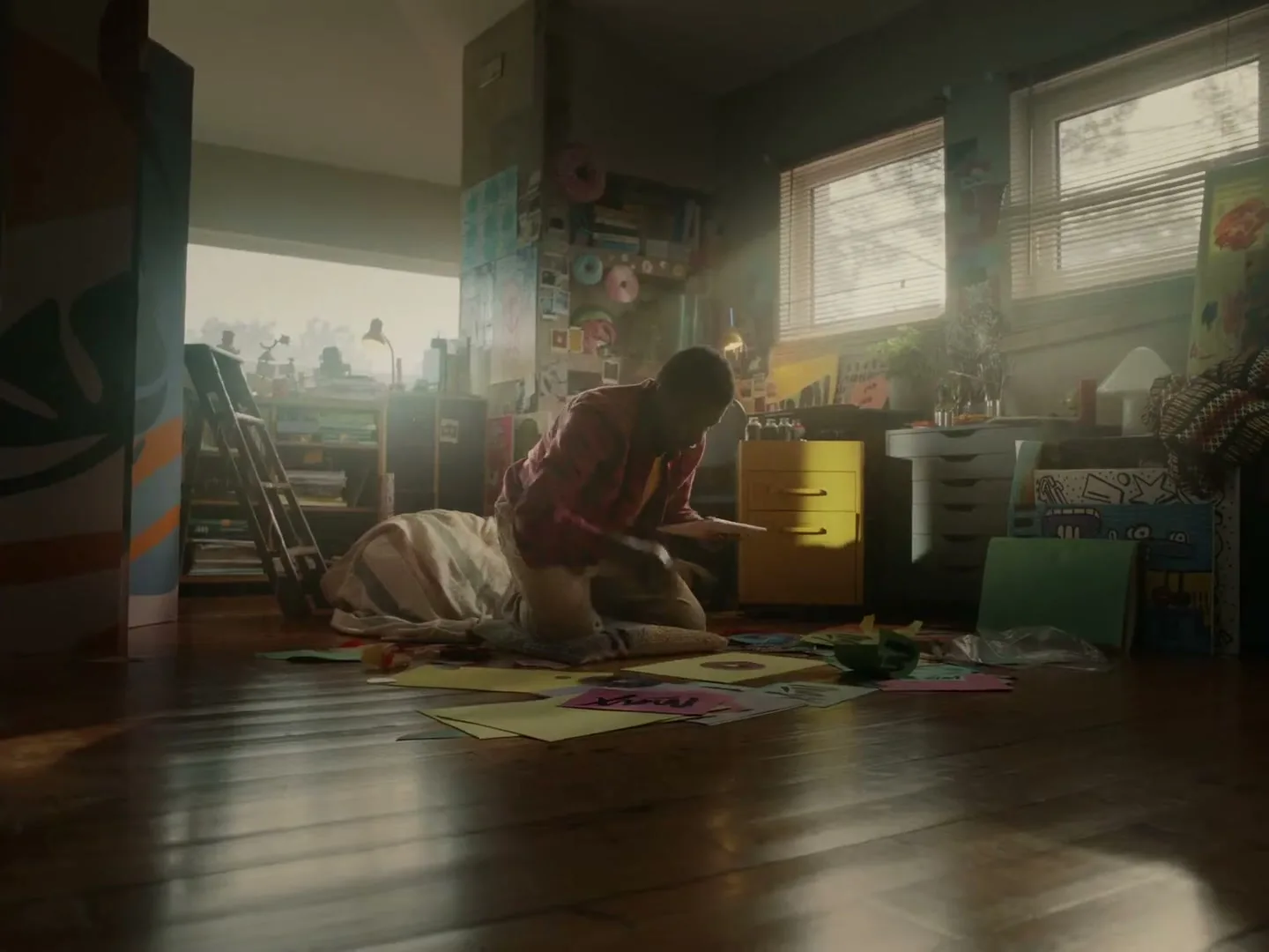 A young Black man in a pink plaid shirt kneels on a wooden floor, surrounded by colorful papers, working on an art project in a sunlit, cluttered room.