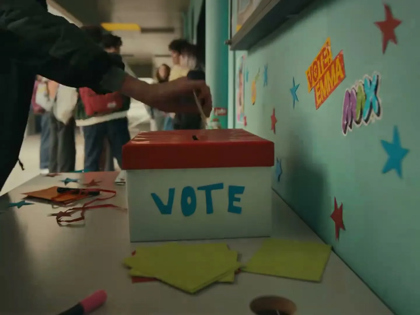A student's hand drops a ballot into a homemade "VOTE" box on a table, surrounded by other students in a school hallway.