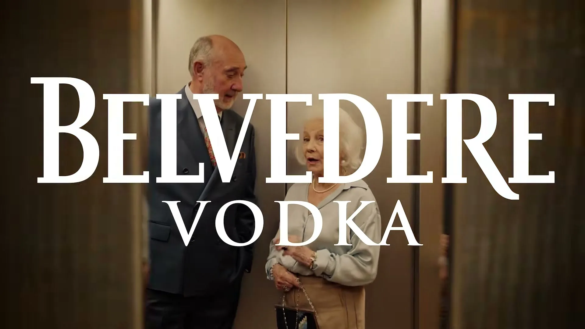 An older man and woman in formal attire are standing in an elevator, talking, as the doors close on them.