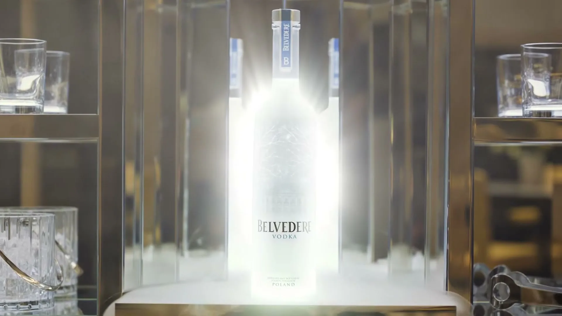 A glowing bottle of Belvedere Vodka is displayed in a mirrored cabinet, surrounded by glassware and bar tools.