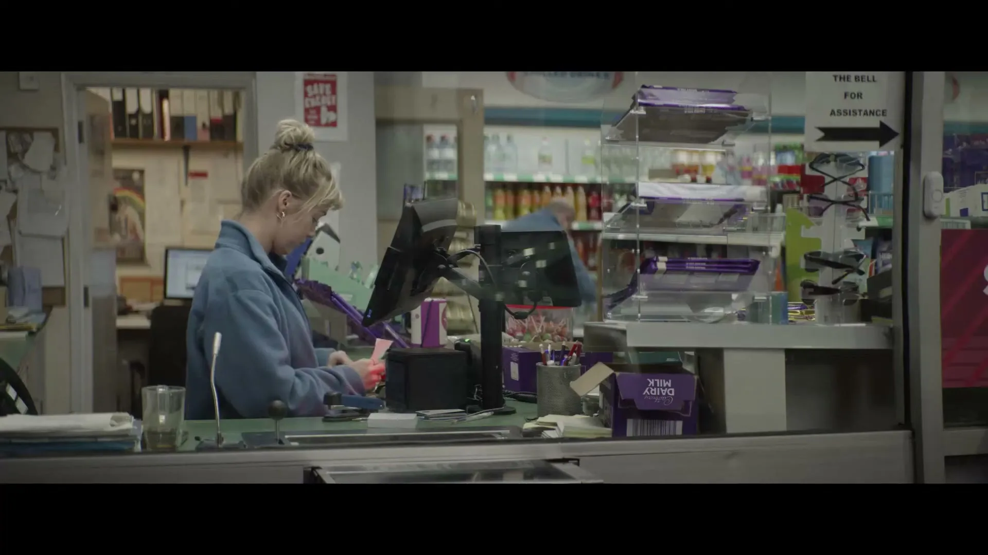 A young woman with blonde hair in a bun, wearing a blue fleece, works at a counter in a garage shop during the day.