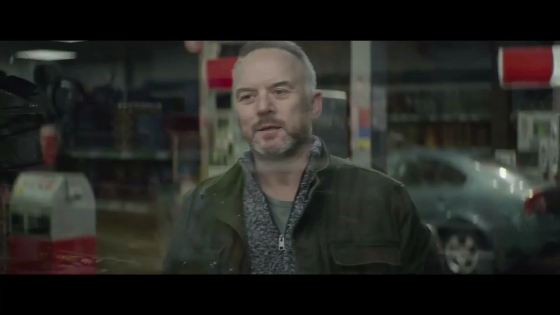 A middle-aged man with a beard and a green jacket looks around a gas station convenience store at night, seen through a reflection.
