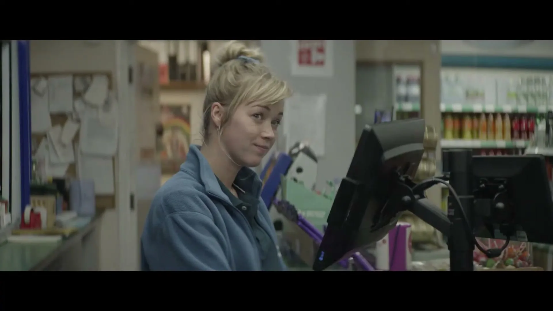 A young female cashier with blonde hair tied in a bun, wearing a blue fleece, looks up from her register and smiles at a customer in a convenience store.