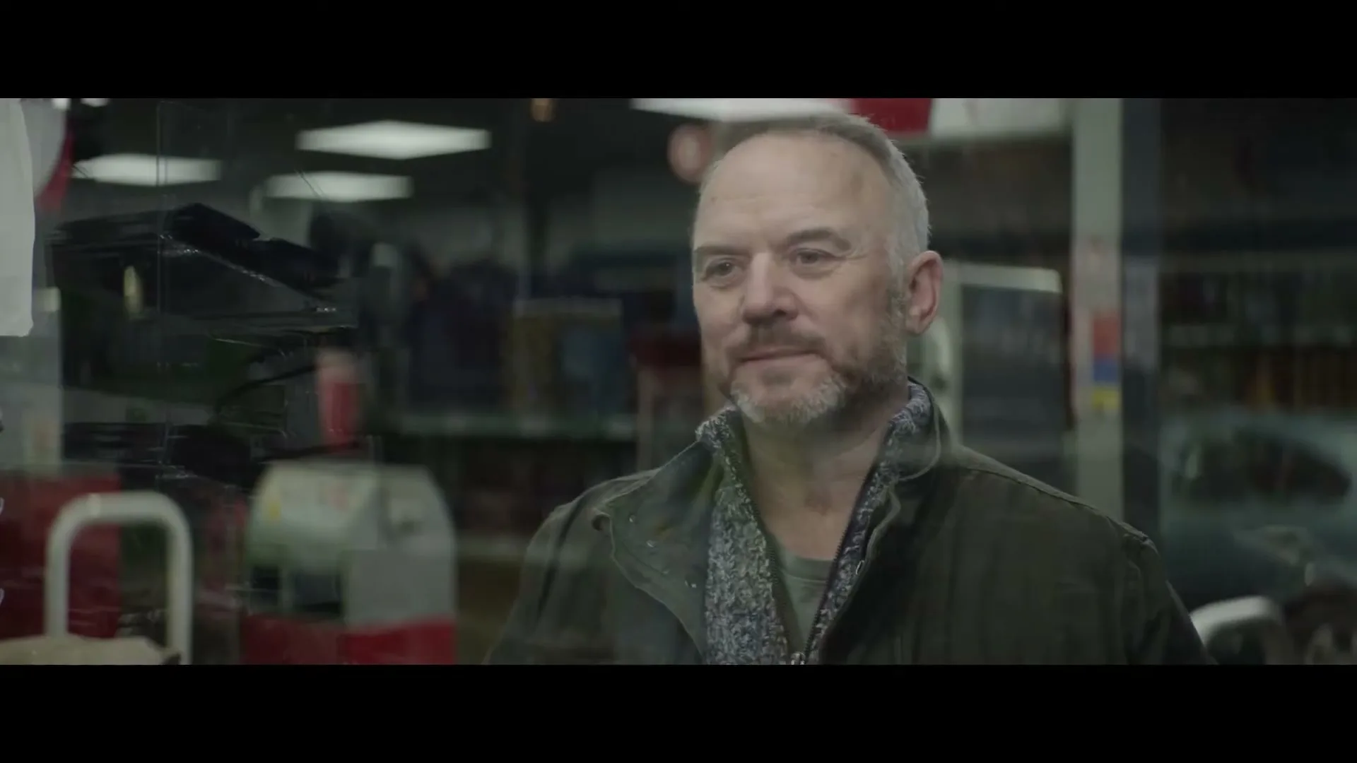 A middle-aged man with a beard, wearing a jacket, looks through a shop window at a chocolate bar, then turns to the side and smiles.
