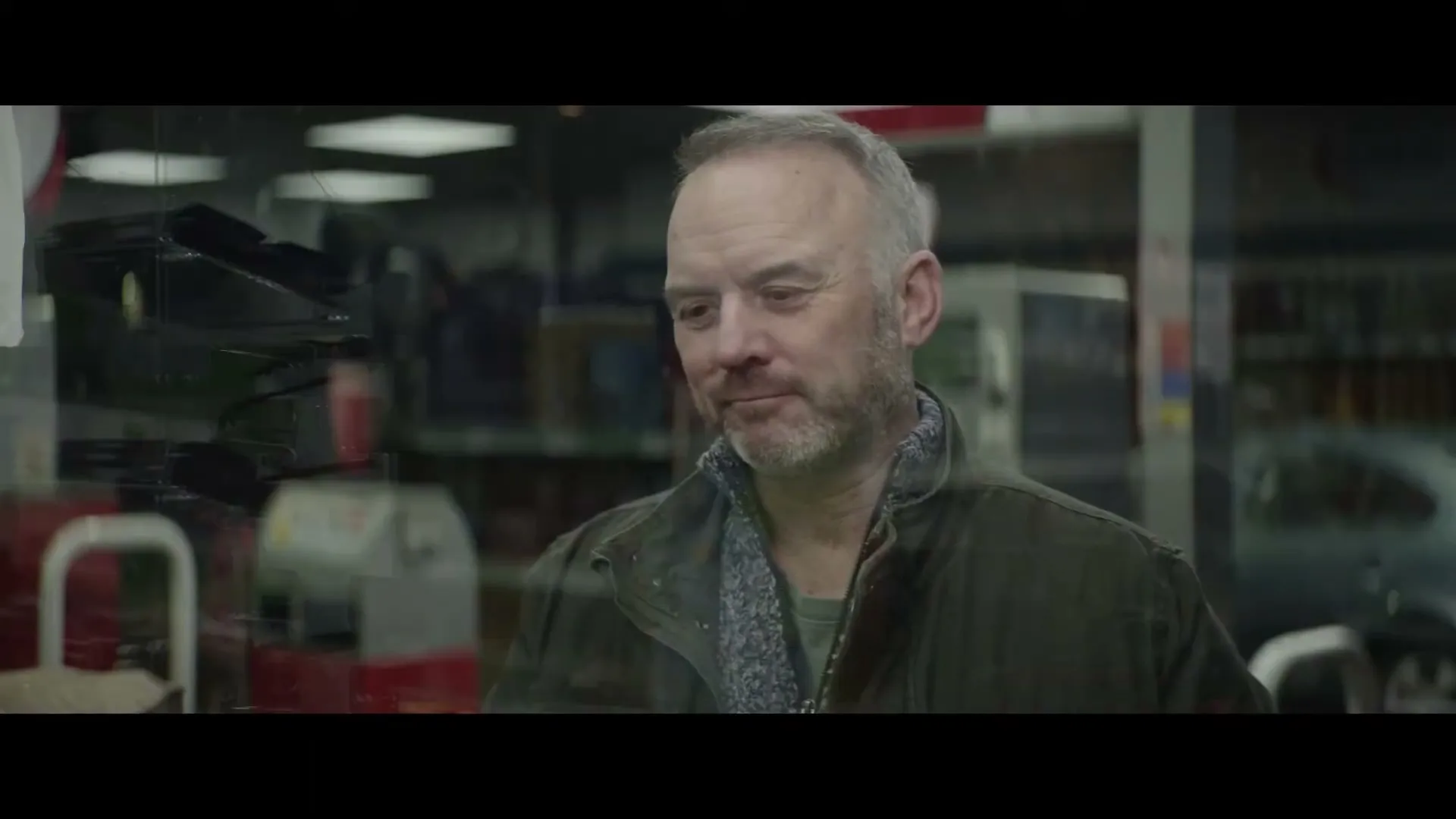 A middle-aged man with a beard and a green jacket looks through a window, smiling slightly, in a garage or auto shop.