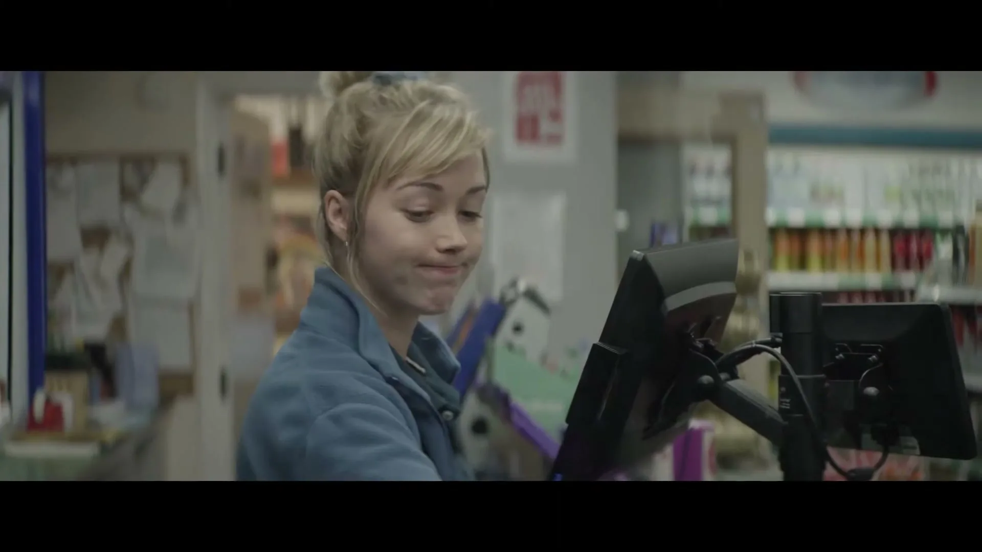 A young female cashier with blonde hair in a blue uniform looks around and then down at the register in a convenience store.