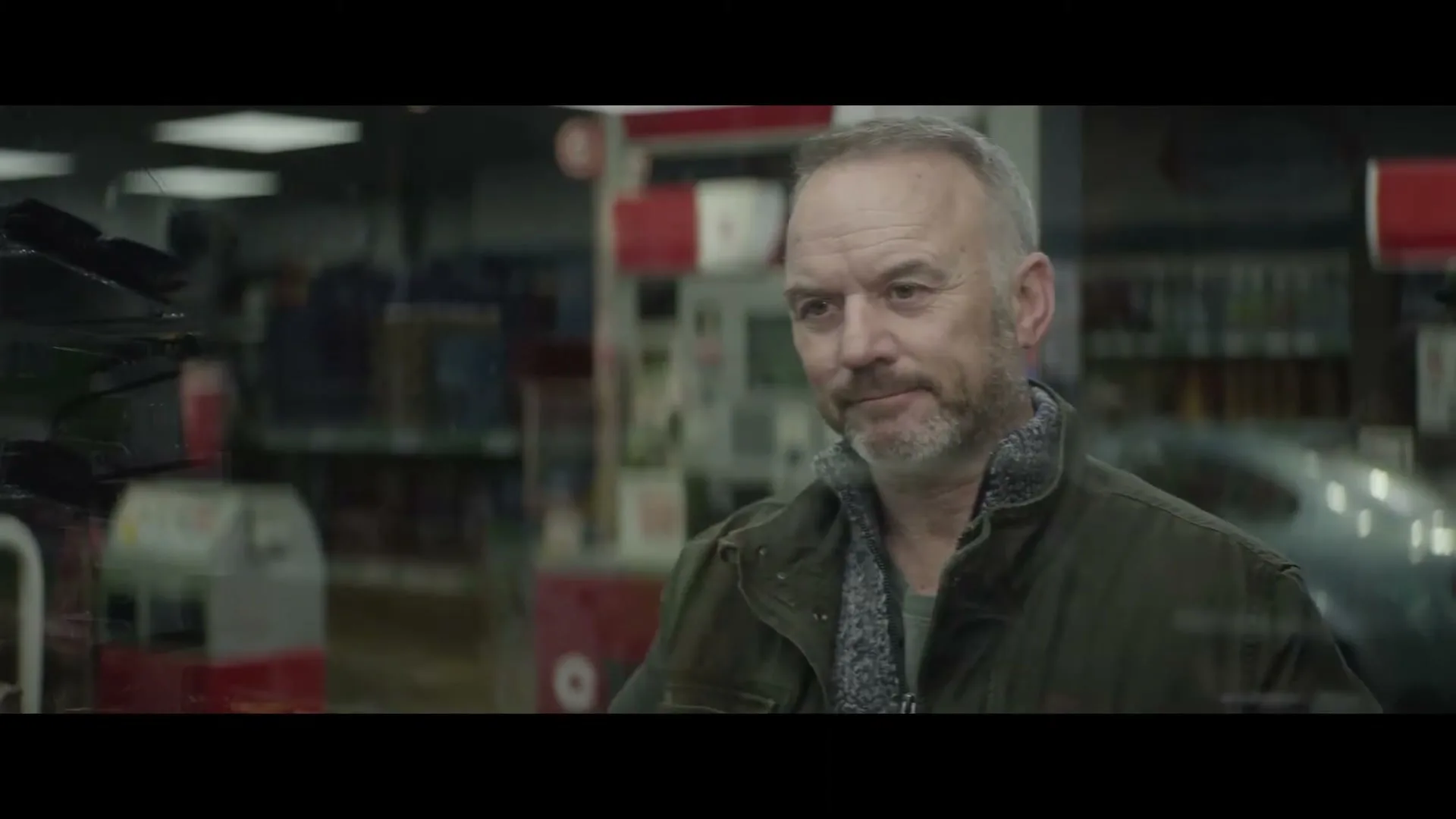 A middle-aged man with a beard, wearing a jacket, smiles and then turns to walk away inside a gas station convenience store.
