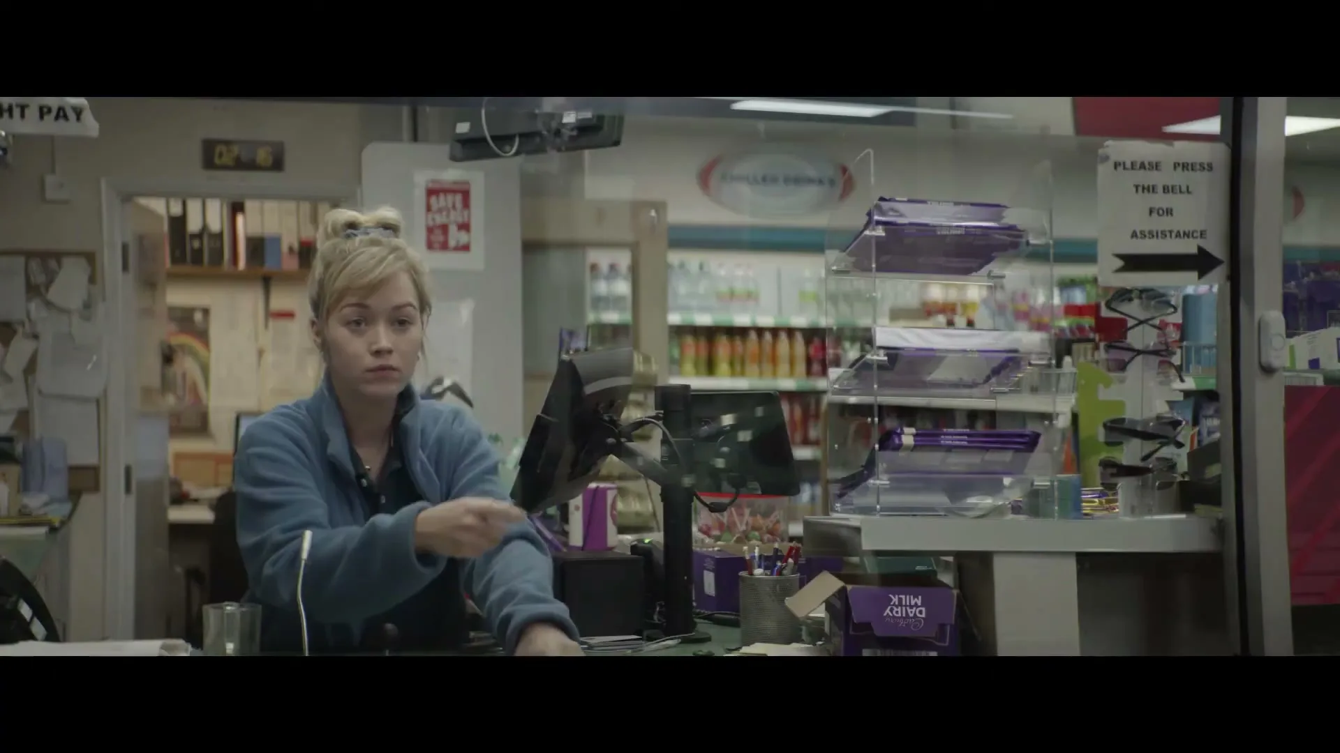 A young female cashier with blonde hair in a bun, wearing a blue fleece, scans a Cadbury Dairy Milk chocolate bar at a convenience store till.