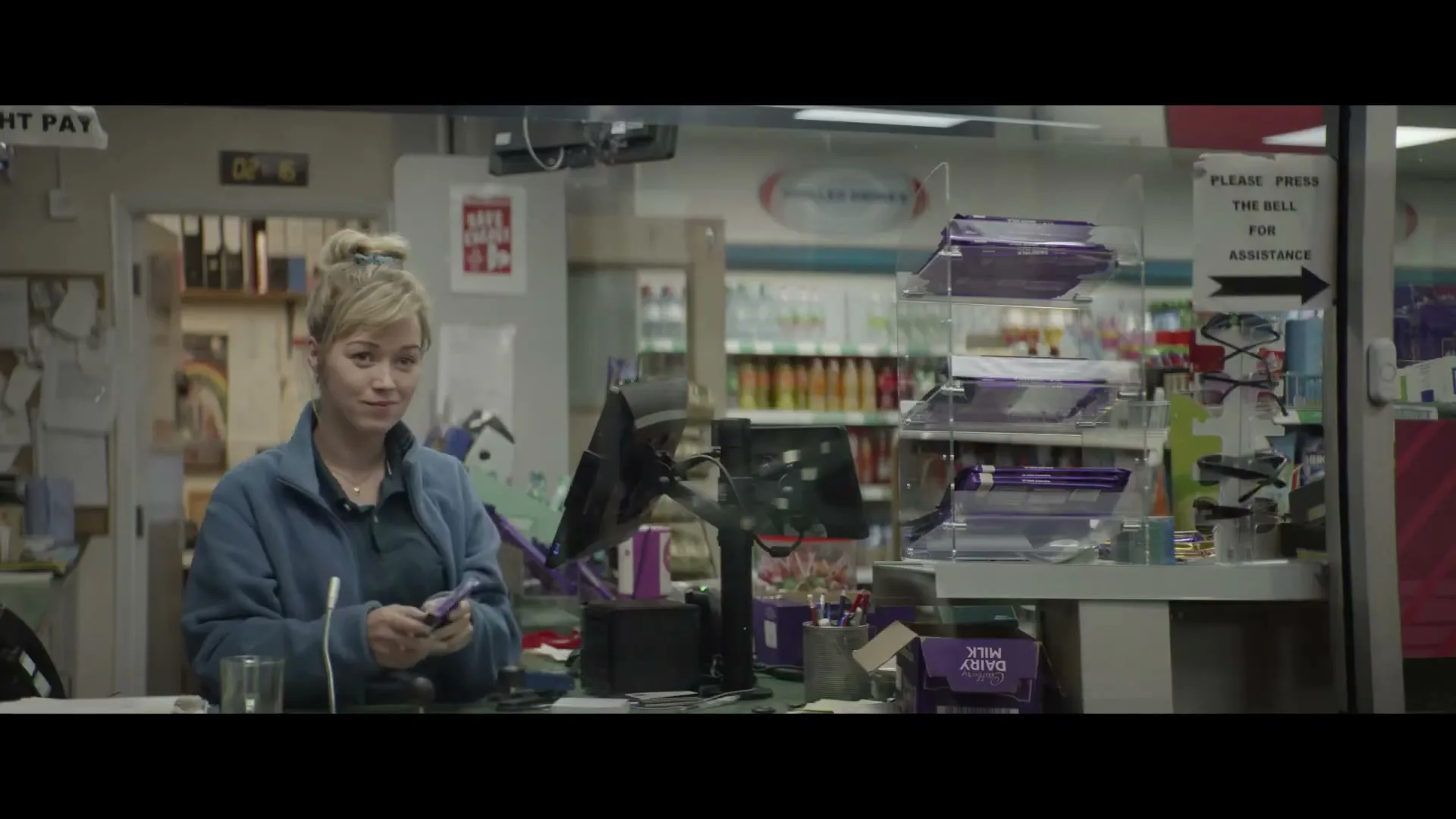 A female cashier with blonde hair in a blue fleece jacket smiles and then looks down at a Cadbury chocolate bar at a gas station convenience store counter.